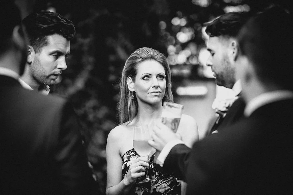 Group of four people at a formal outdoor event, with woman in the center holding a glass of wine, surrounded by three men in tuxedos.