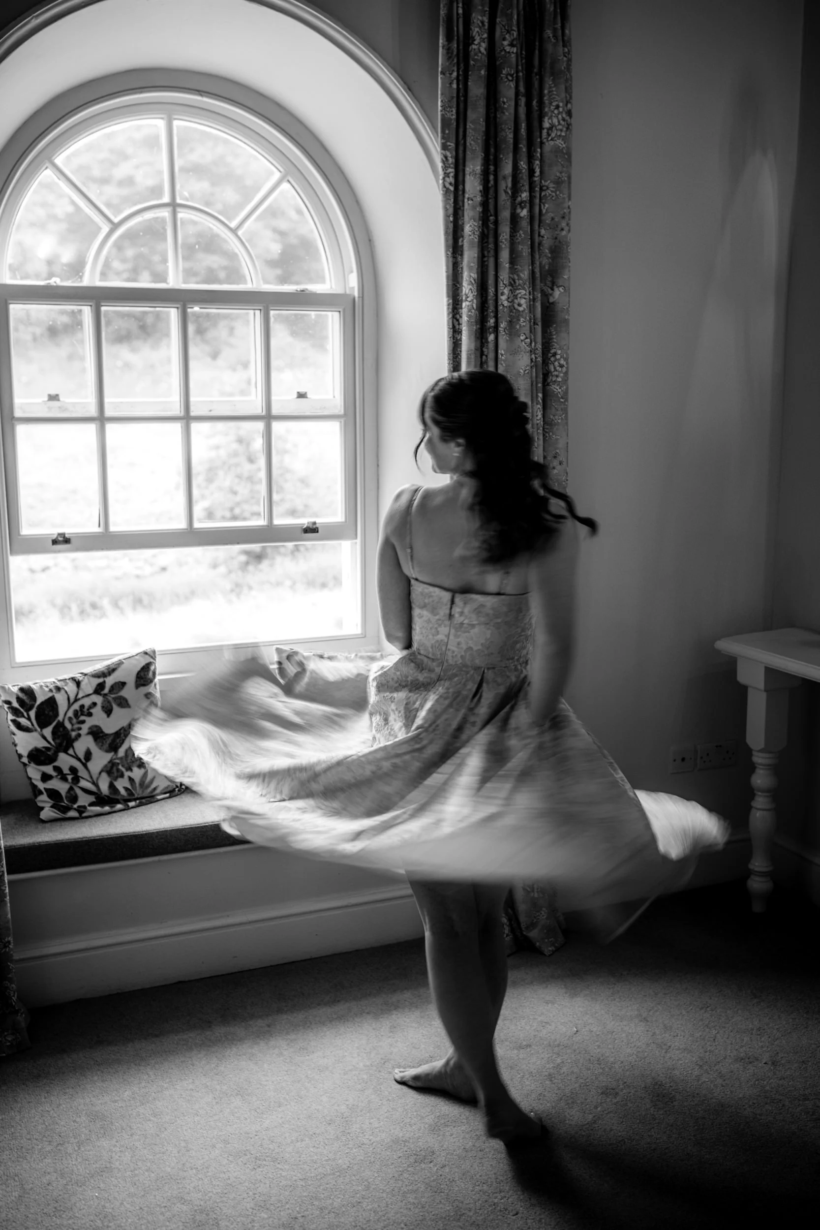 A woman with dark hair, wearing a dress, is standing in front of a large, ornate window with curved top and multiple panes, in a room with floral curtains and a small table. She appears to be adjusting a bedspread or blanket on a bed with decorative pillows.