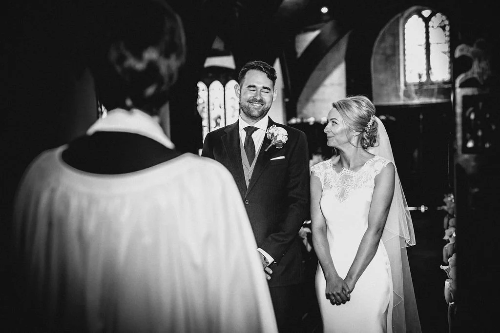 A black and white photo of a wedding ceremony inside a church. The bride and groom are smiling, standing before an officiant, with the groom in a suit and the bride in a wedding dress and veil.