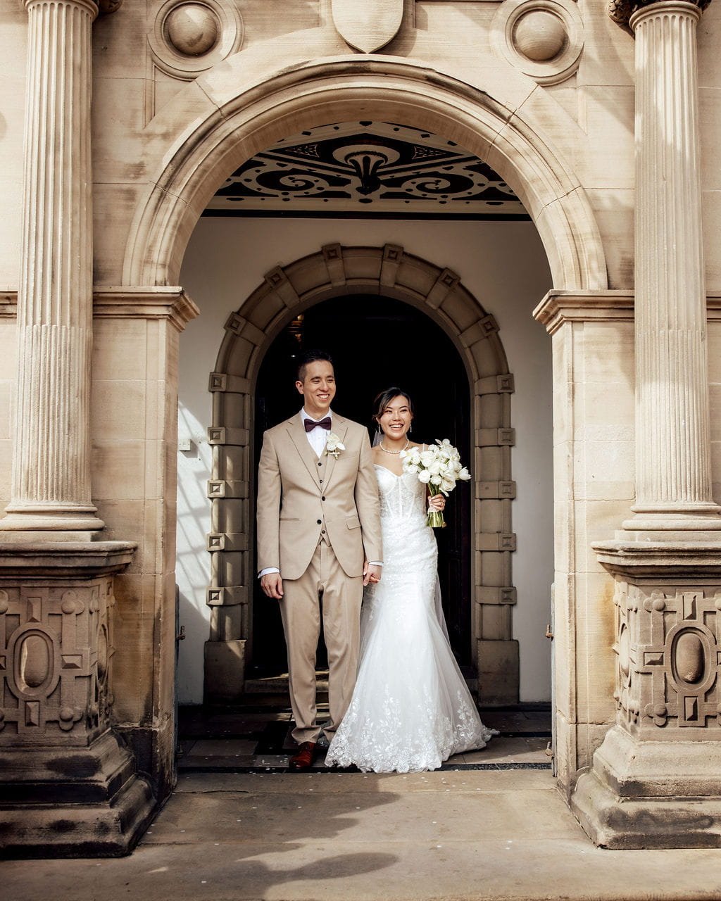 A newlywed couple standing under an ornate stone archway, smiling. The bride, holding a bouquet of white flowers, is wearing a white wedding dress. The groom is dressed in a beige suit with a bow tie.