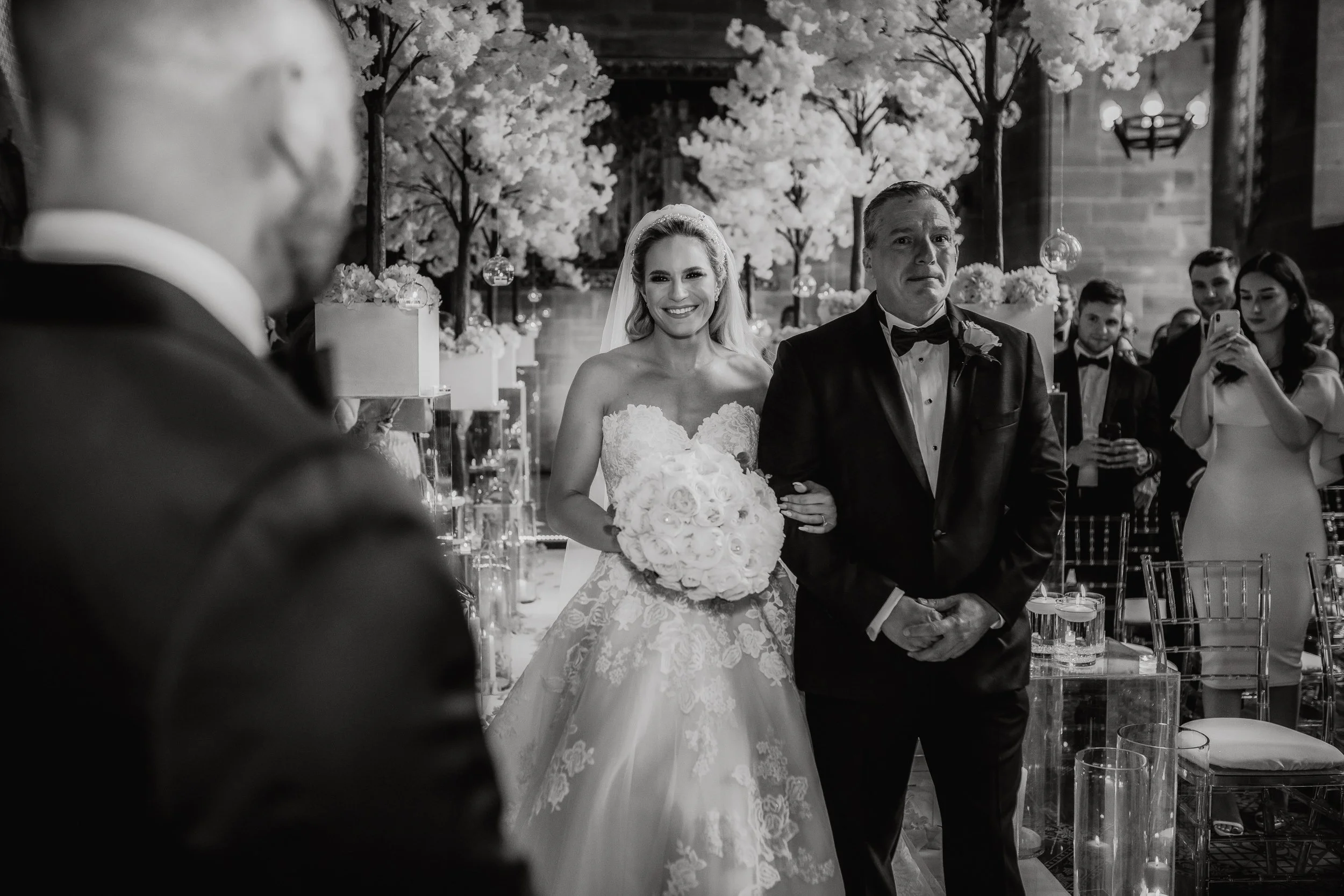 A bride in a lace wedding gown holding a bouquet of roses, being escorted down the aisle by her father in a tuxedo, during a wedding ceremony in a decorated venue with floral arrangements and guests watching.
