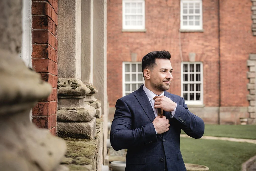 A man in a blue suit adjusting his tie outside a brick building.