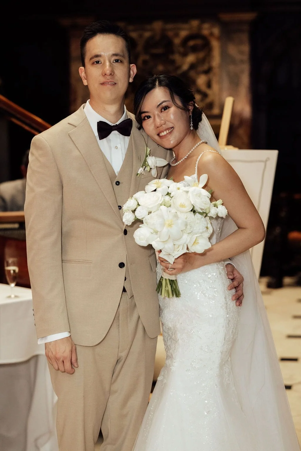 A newlywed couple posing at their wedding reception, with the bride holding a white floral bouquet and wearing a white wedding gown, and the groom in a beige suit with a black bow tie.