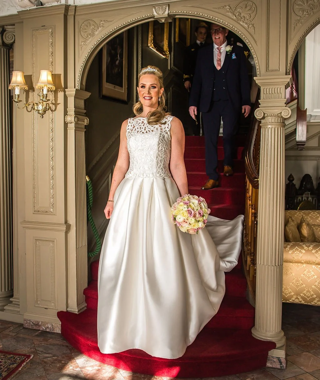 A bride in a white wedding dress holding a bouquet, standing on a staircase with red carpet, smiling as she walks down, with a man in a suit and tie smiling behind her.