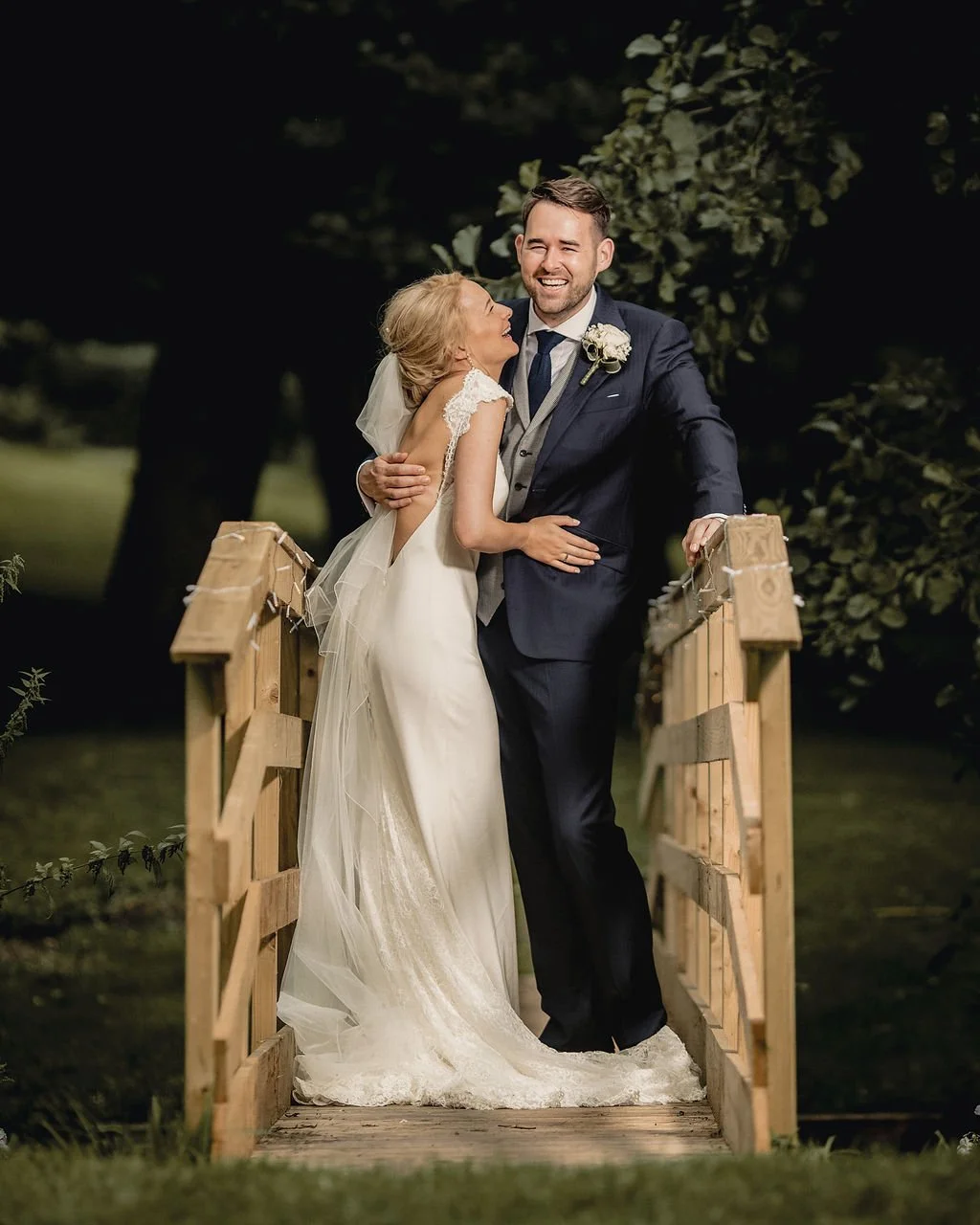 A bride and groom embracing on a wooden bridge outdoors at night, smiling and happy.