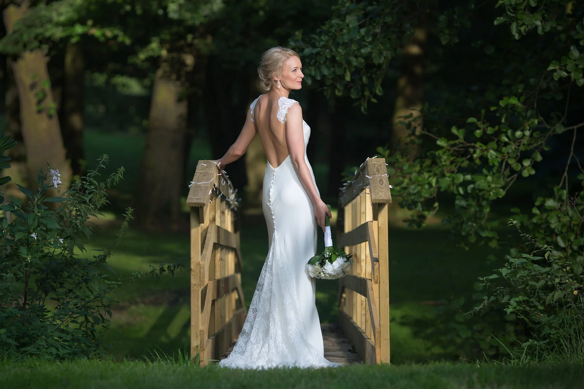 A bride in a white wedding dress with lace details on the shoulders standing on a small wooden bridge in a lush garden, holding a bouquet of white flowers, with trees and foliage in the background.