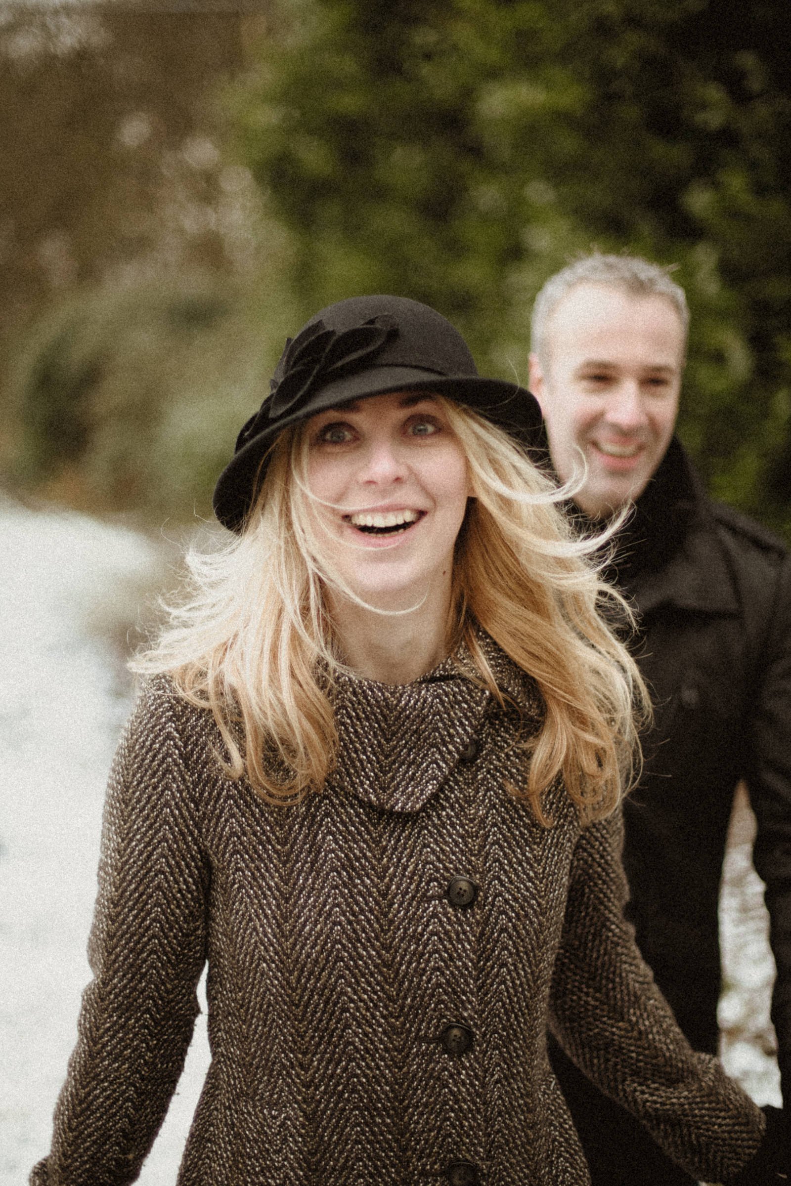 A woman with long blonde hair wearing a black hat and a brown herringbone coat, smiling outdoors with a man in a black jacket in the background.