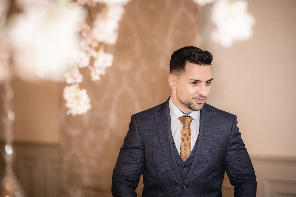 A man in a dark, checkered suit and a white shirt, with a light brown tie, standing indoors with floral decorations and a blurred background.