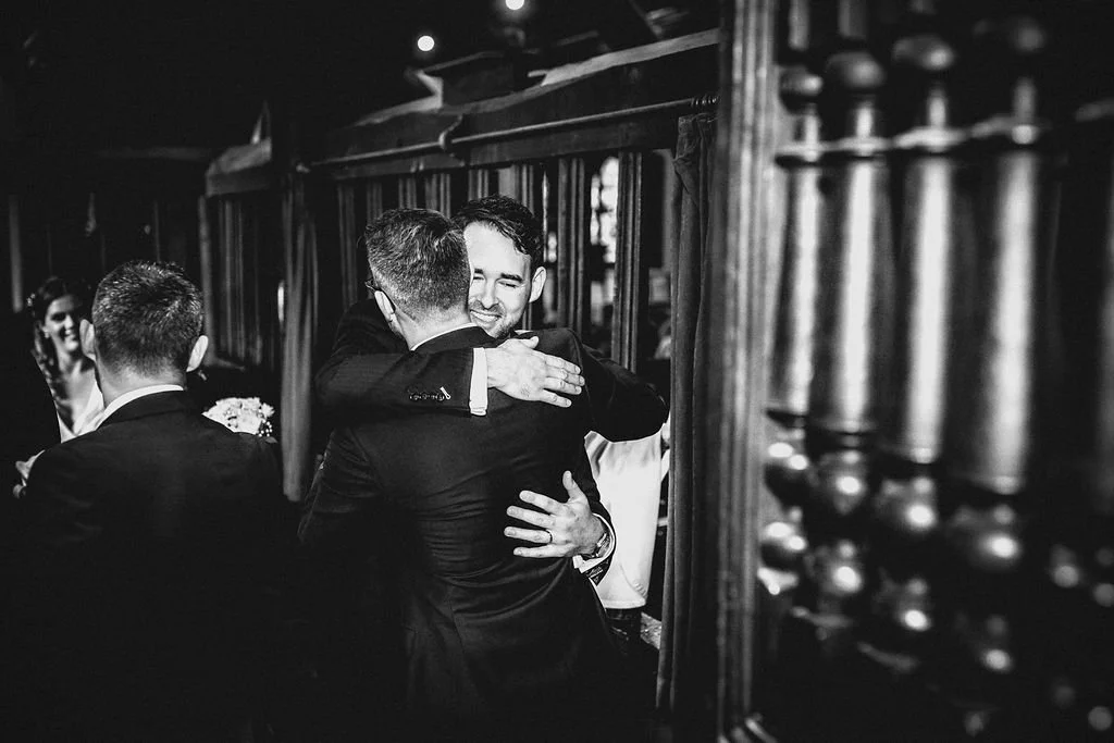 Two men in suits hugging and smiling at a wedding reception, with other guests in the background.