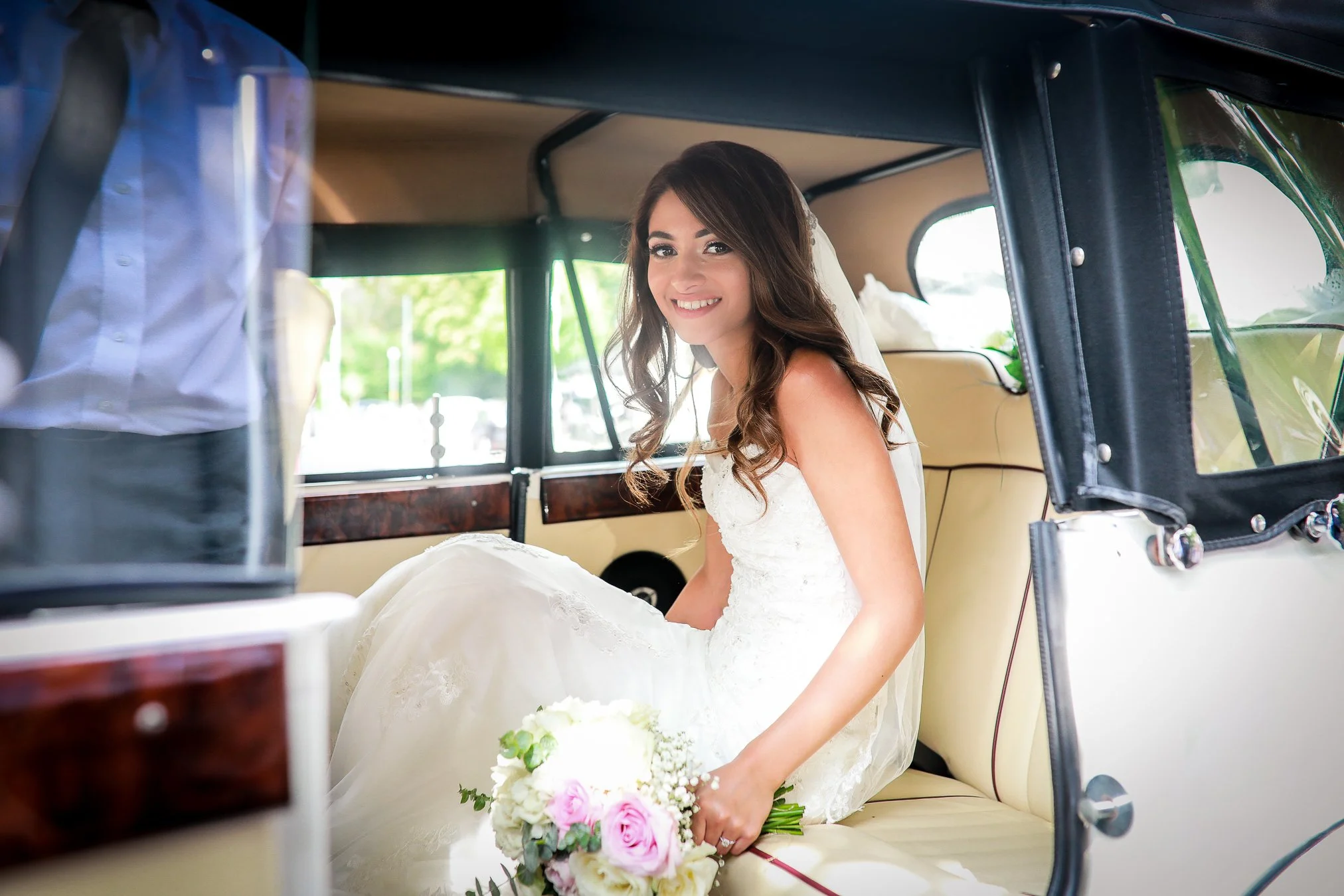 Beautiful young bride wearing a white wedding dress and holding a pink and white bouquet of flowers, smiles as she is about to exit her wedding car.