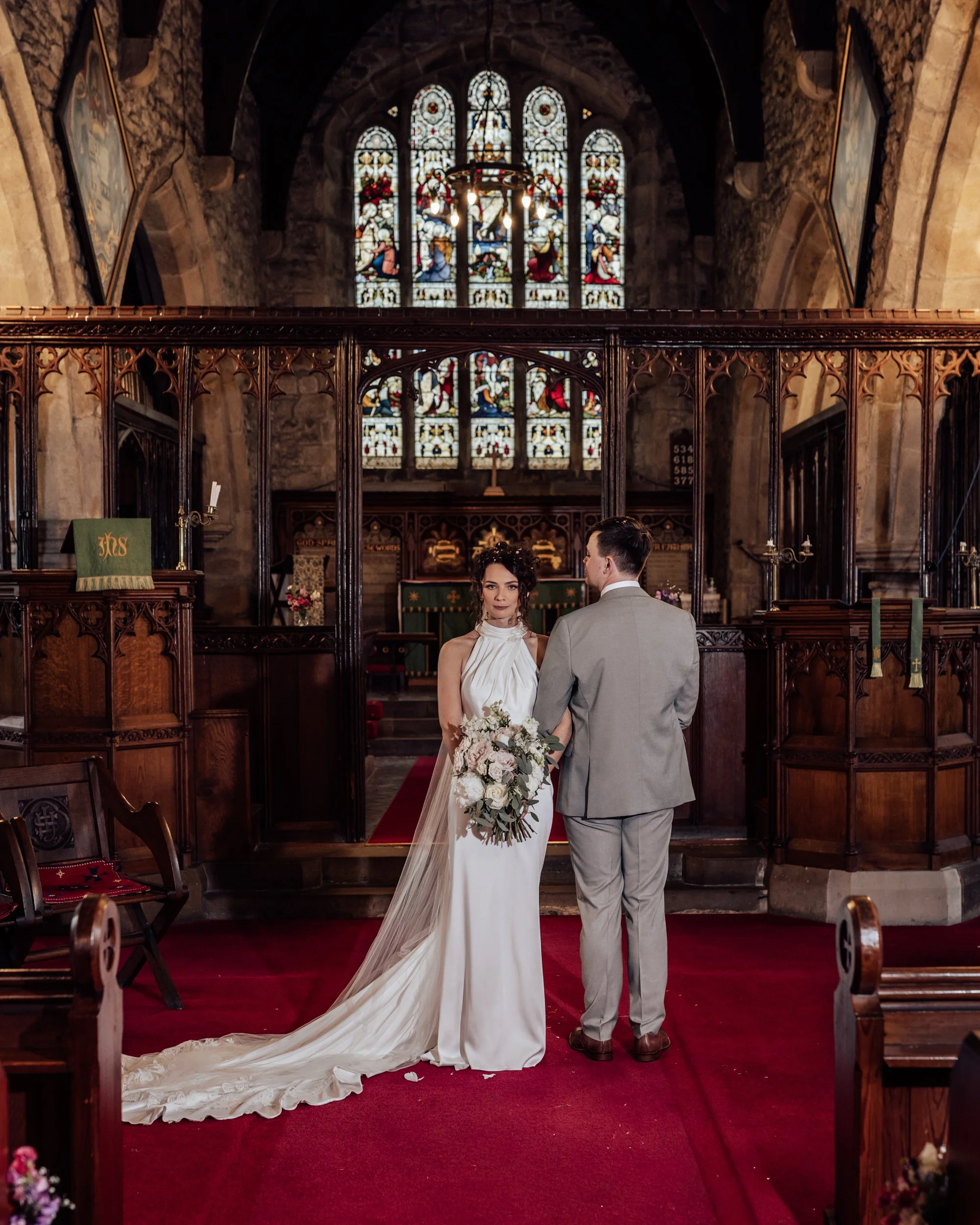 A bride and groom standing inside a church during a wedding ceremony, with stained glass windows and wooden pews in the background.