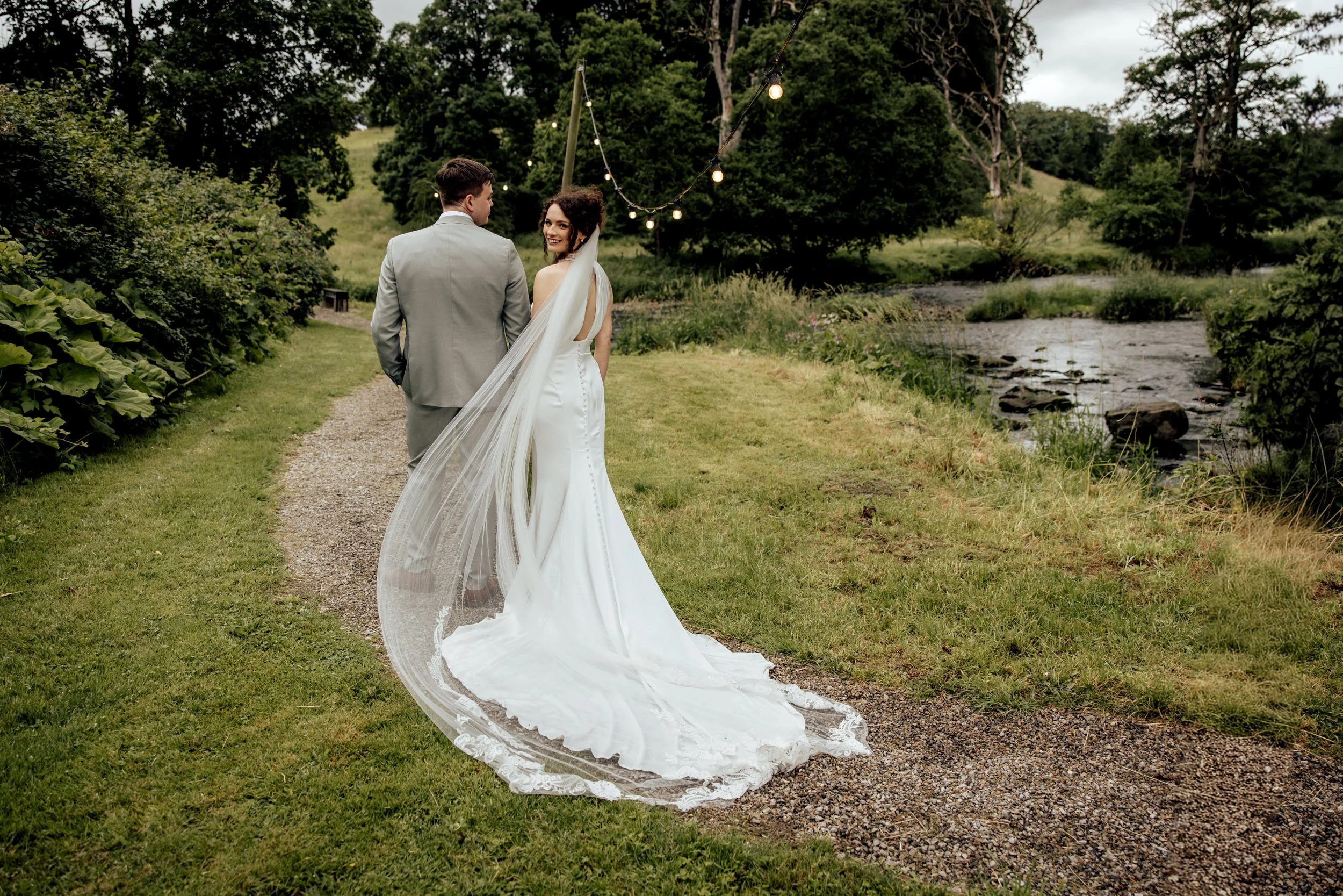 Bride and groom walking on a gravel path beside a stream in a lush, green outdoor setting, with string lights overhead during daytime.