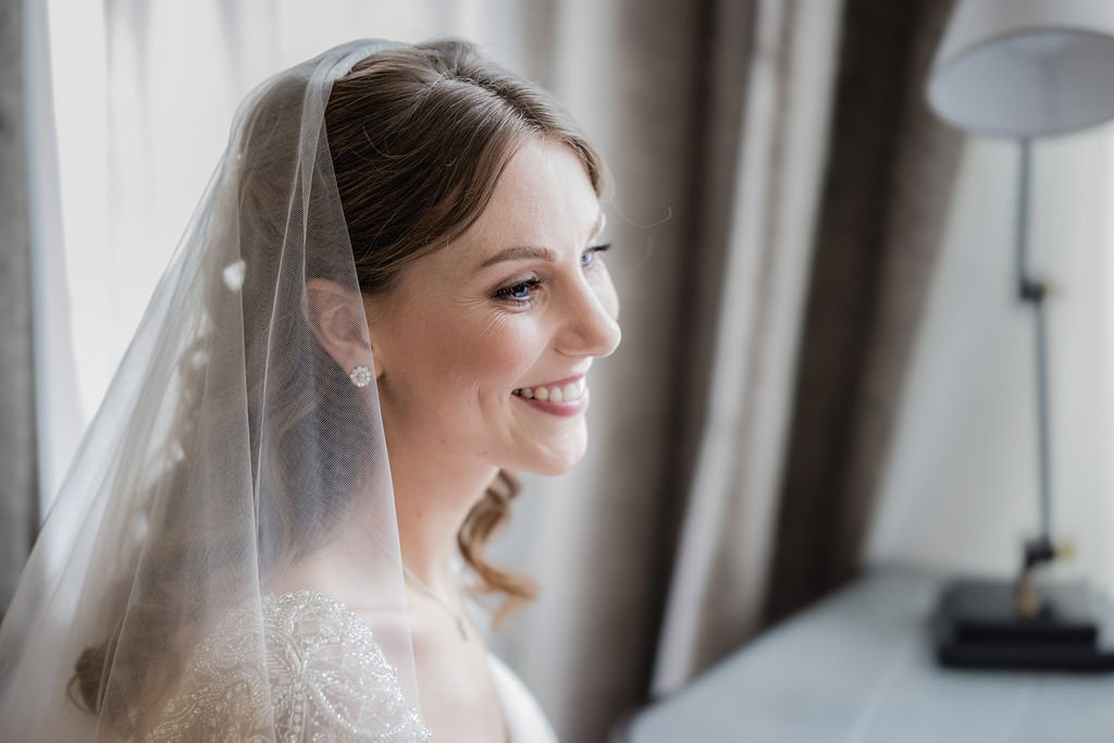 A bride with a veil and wedding earrings, smiling, looking out a window in a softly lit room.