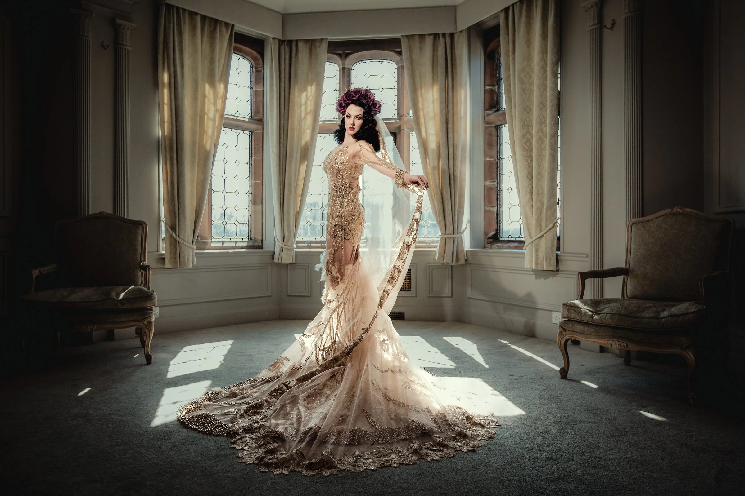 A woman in an elegant, beige, lace wedding dress with a long train, standing in a sunlit, vintage-style room with large windows and cream-colored curtains.