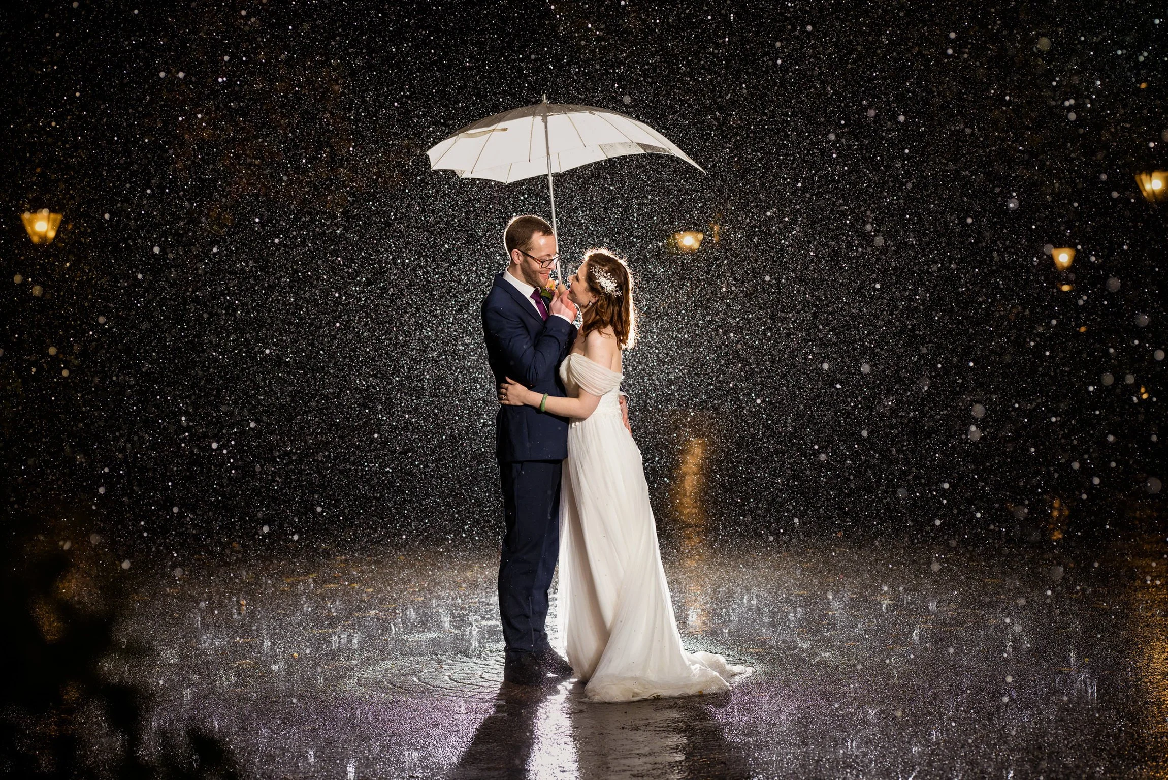 A bride and groom stand close together under an umbrella in the rain at night, looking into each other's eyes as they embrace.