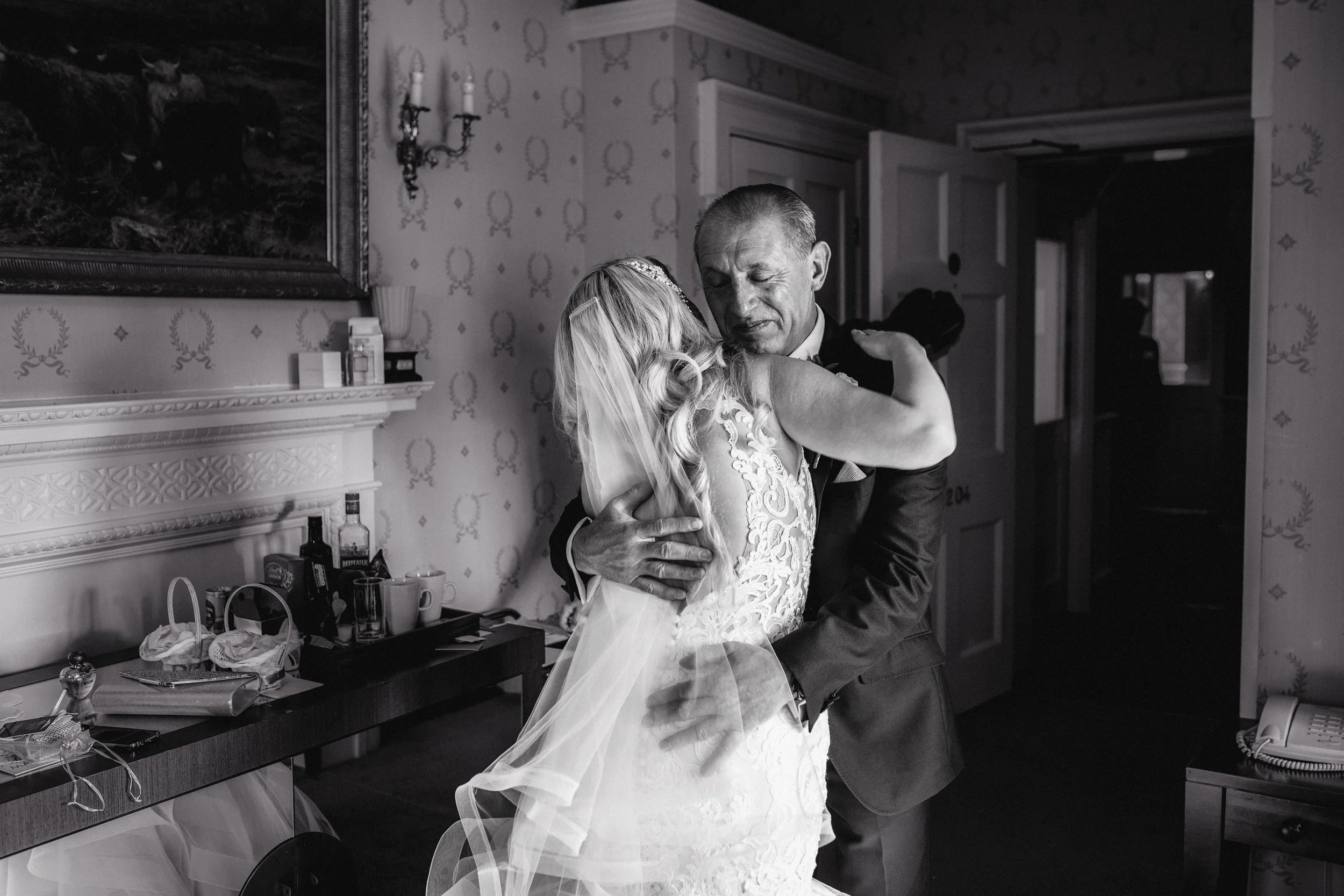 A black-and-white photo of a bride and an older man in an embrace, likely during a wedding celebration, inside a room with patterned wallpaper and a fireplace with bottles and cups on it.