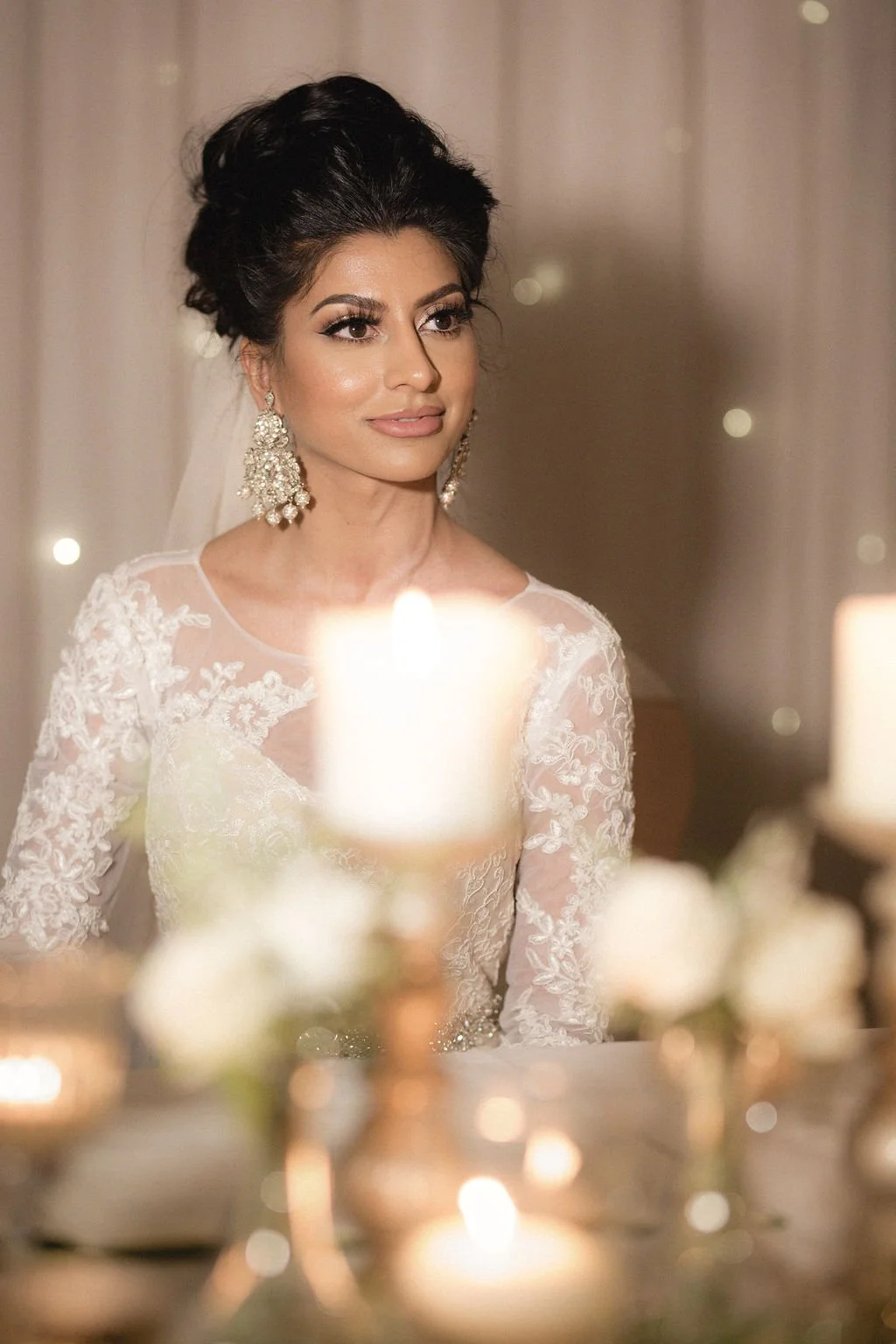 A woman in a wedding dress with lace details and large earrings, standing near candles, with a serene expression on her face.