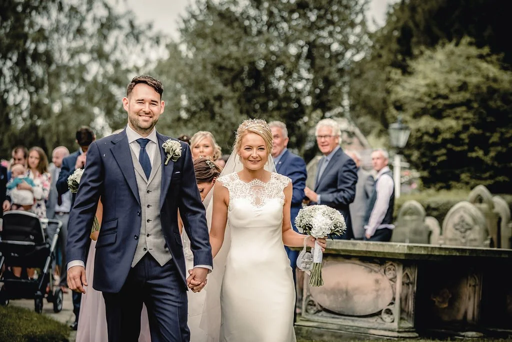 A bride and groom smiling and holding hands during their outdoor wedding ceremony, with guests in the background.