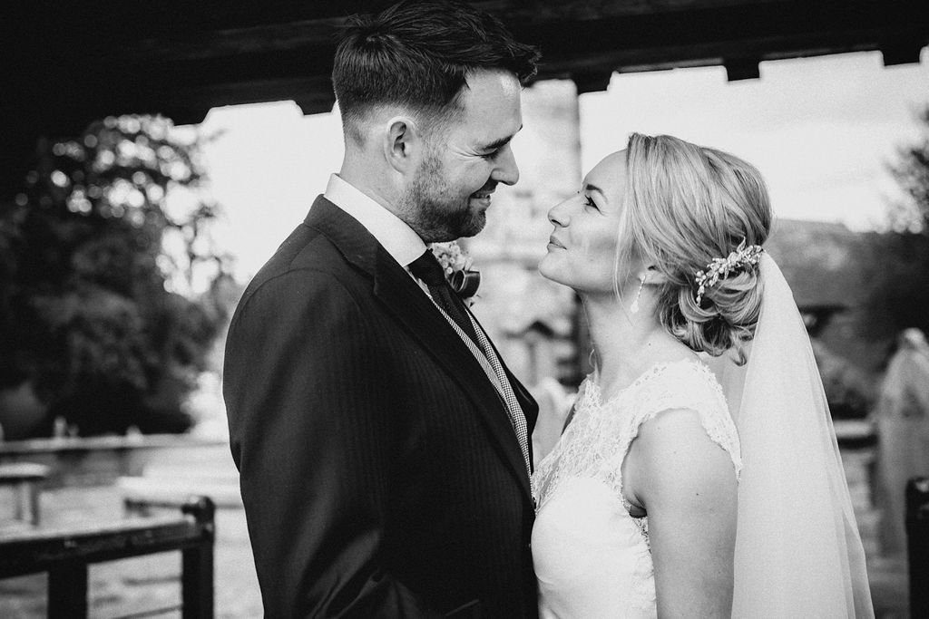 A couple in wedding attire, with the groom smiling and the bride looking into his eyes, under an outdoor pavilion.