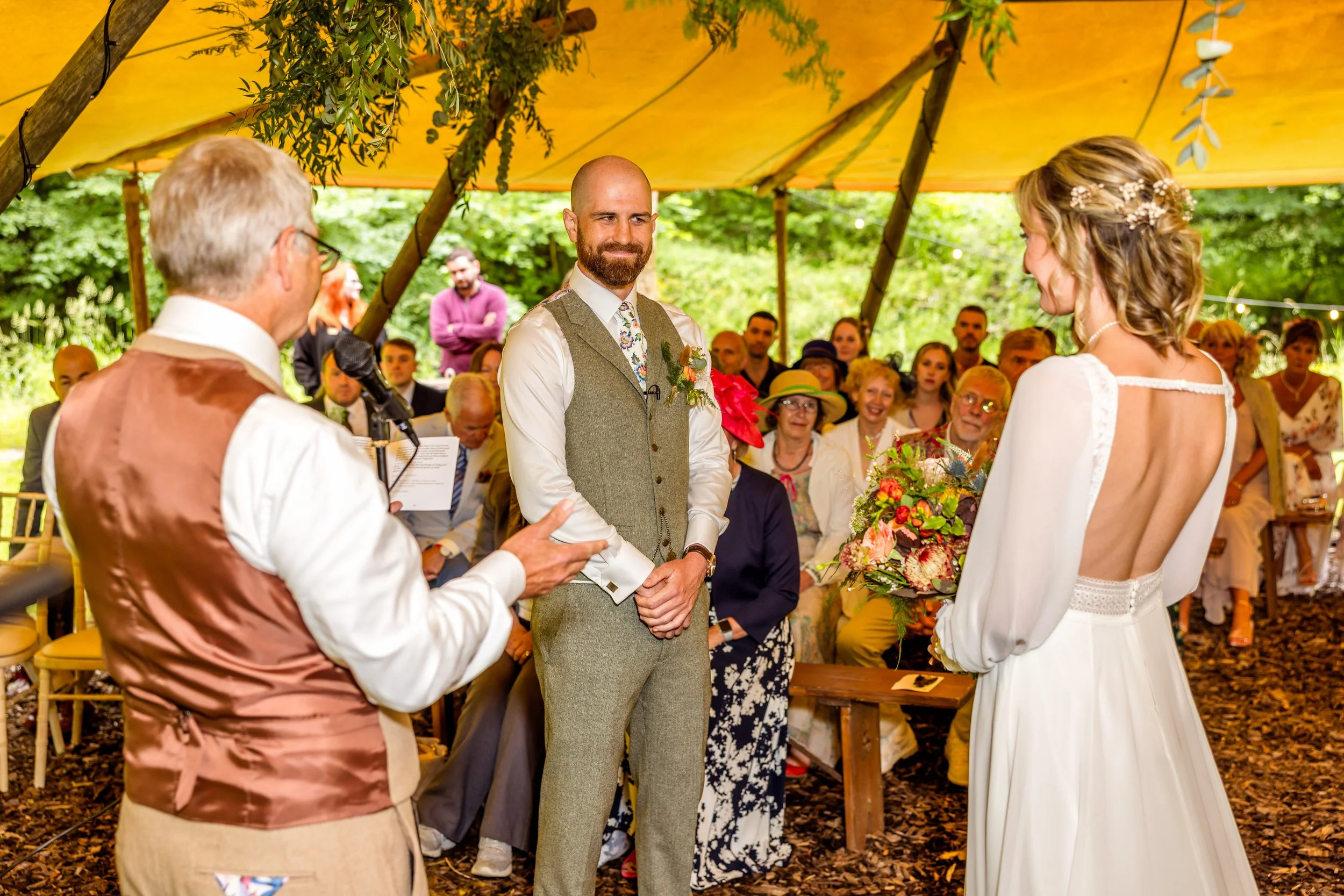 A wedding ceremony taking place outdoors under a yellow canopy with greenery. The bride and groom stand facing each other, with the officiant speaking to them. The bride is holding a colorful bouquet, and the groom is smiling. Guests are seated behin