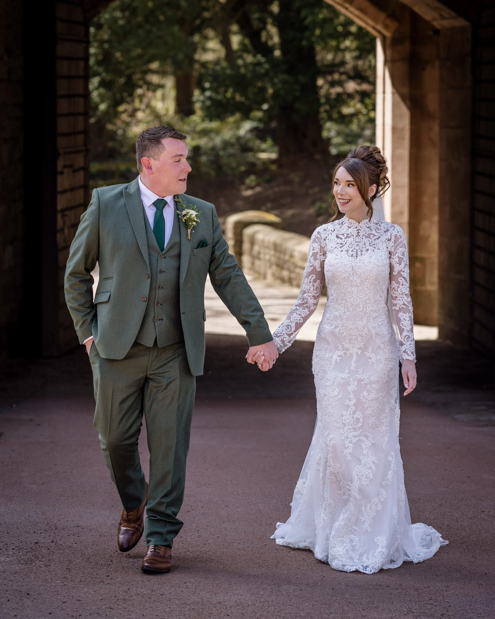 A bride and groom walking hand in hand outdoors, with the groom looking at the bride and the bride smiling, under a brick arch with trees in the background.