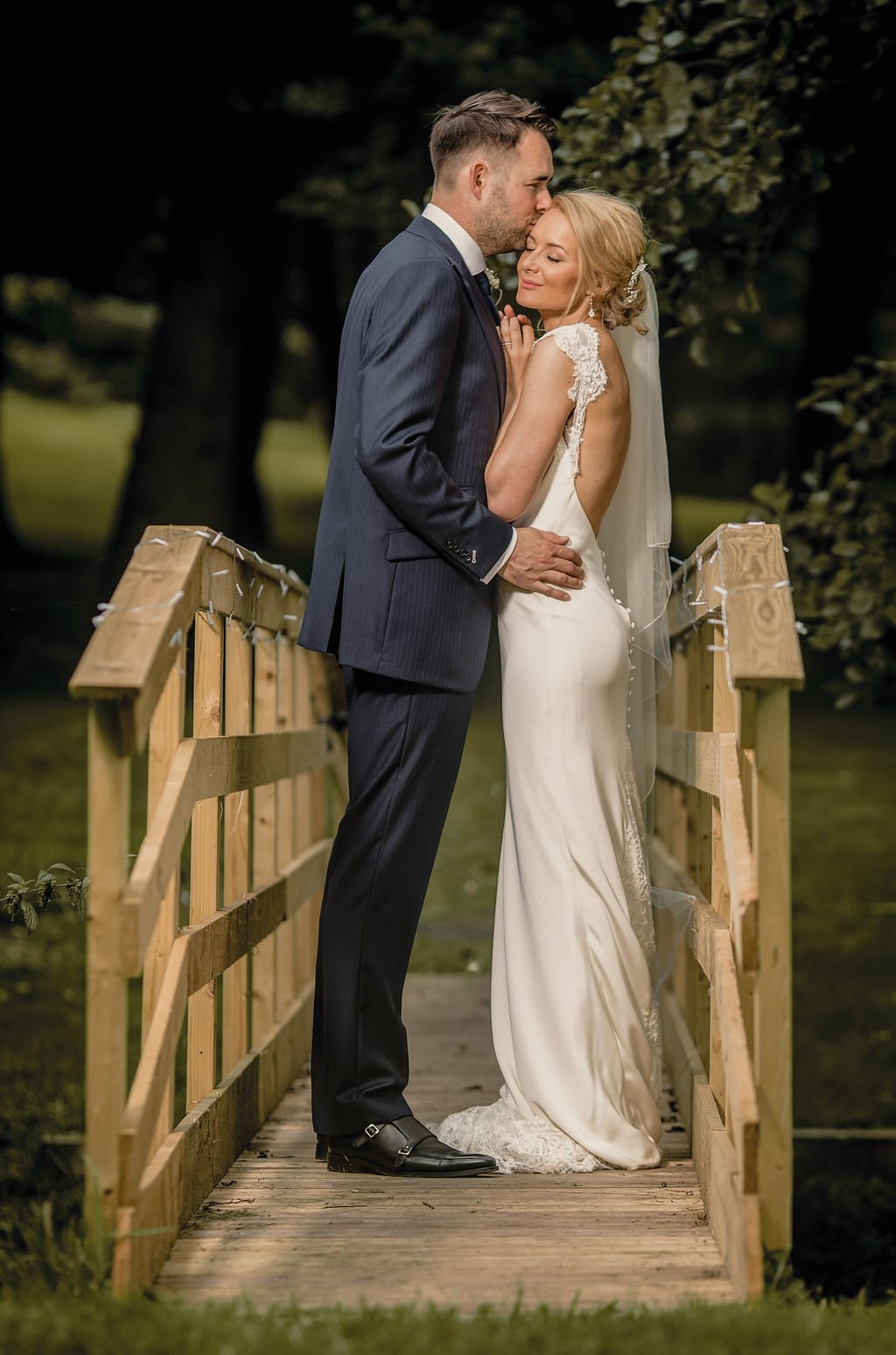 A bride and groom standing on a small wooden bridge outdoors, embracing passionately. The groom, in a dark suit, gently kisses the bride, who is in a white wedding dress, with her eyes closed and her hand on his chest.