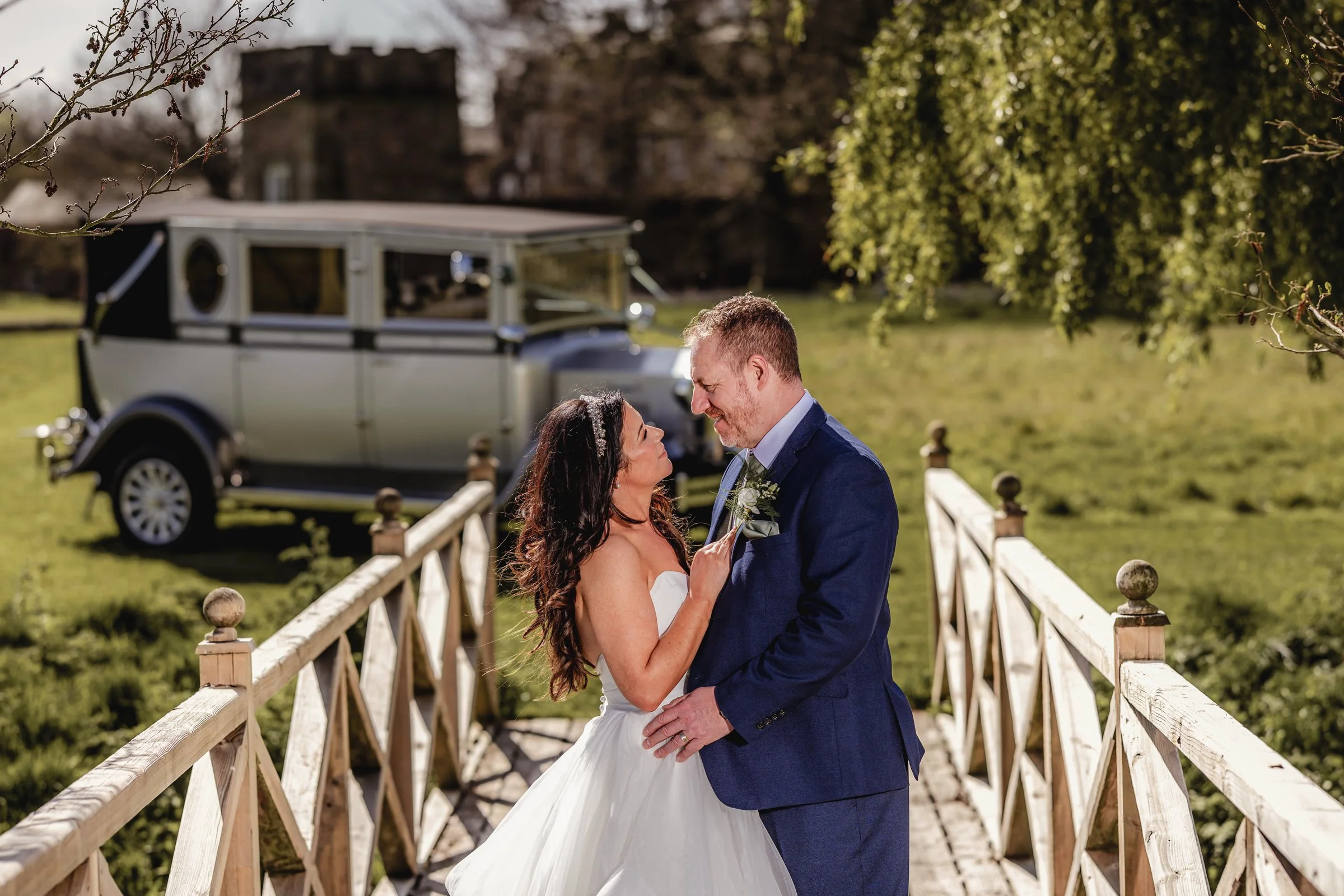 A bride and groom sharing a romantic moment on a small wooden bridge outdoors, with a vintage car in the background.