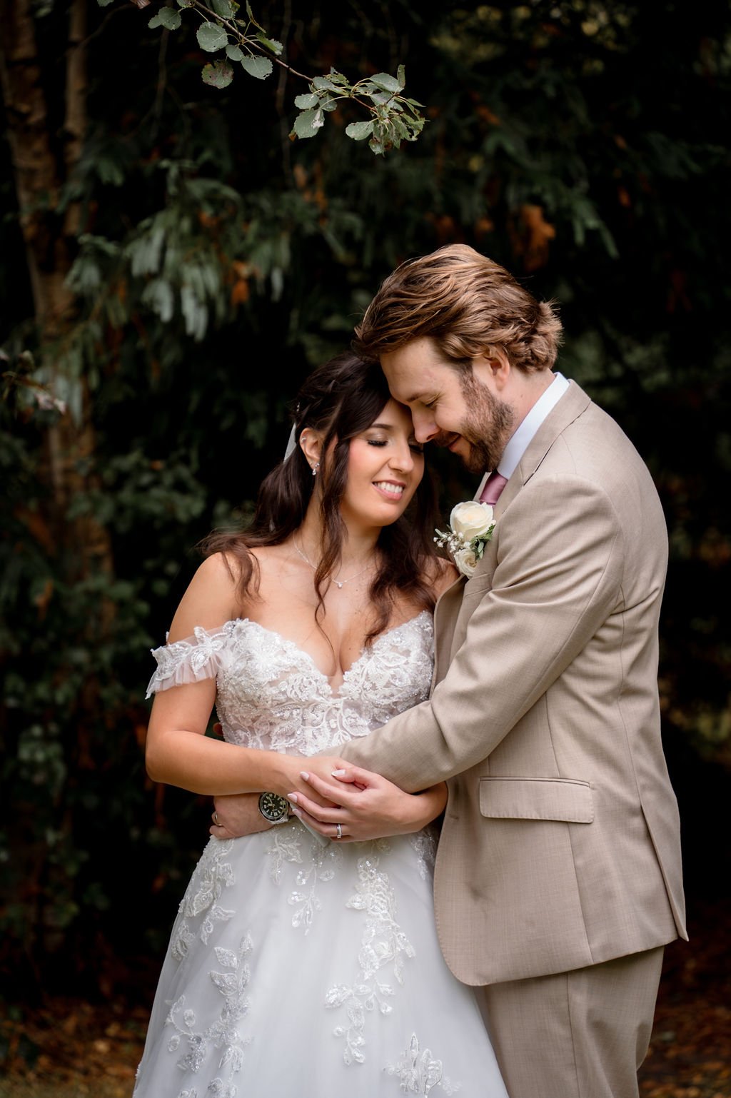 A bride and groom embrace outdoors, with the bride in a lace wedding gown and the groom in a beige suit, against a background of dark green foliage.