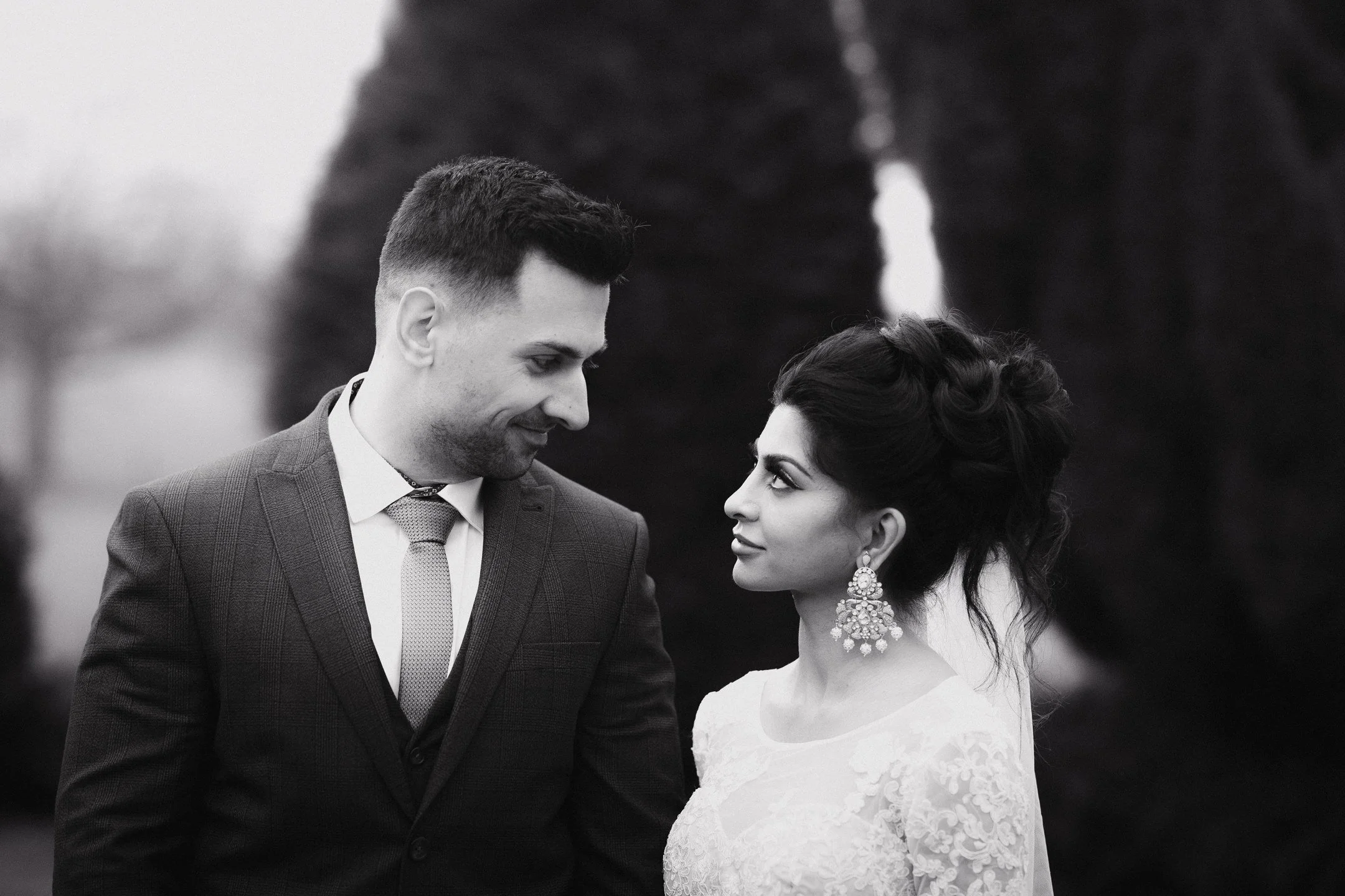 A black and white photograph of a man and woman standing outdoors, gazing into each other's eyes. The man is wearing a suit and the woman is in a lace dress with large earrings, with trees in the background.