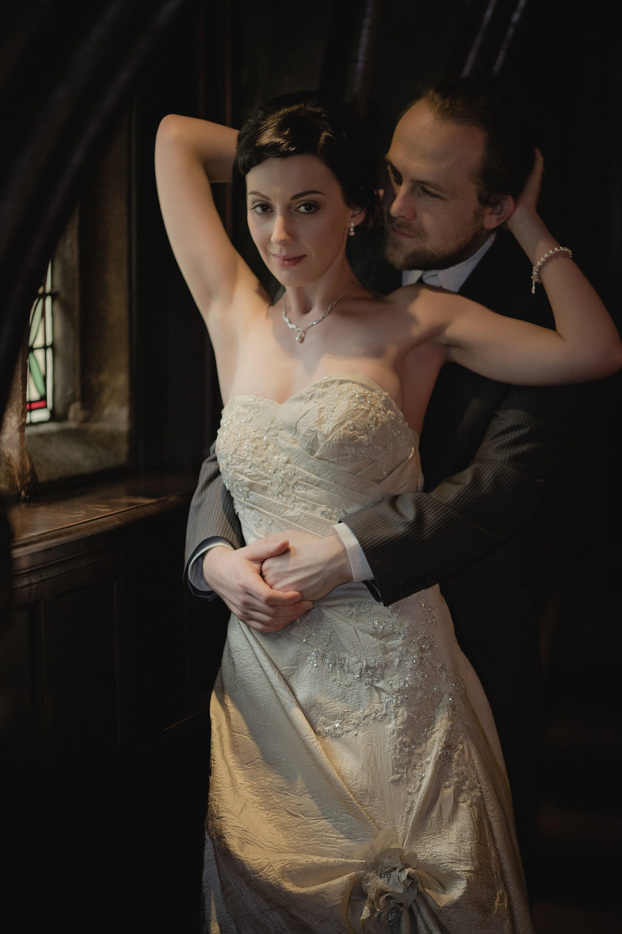 A woman in a wedding dress and a man in a suit embracing inside a dimly lit church, with stained glass windows in the background.