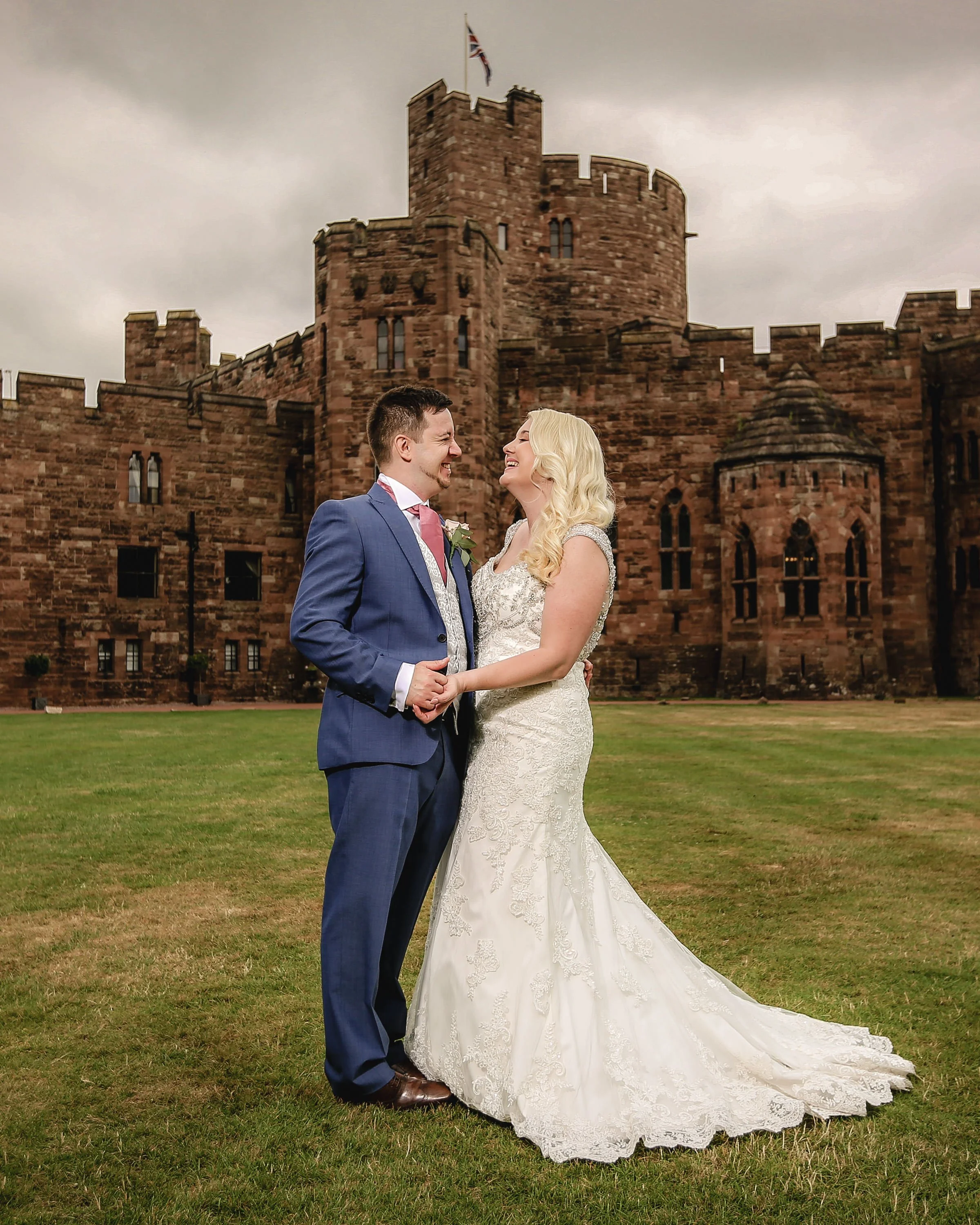 A bride and groom holding hands and smiling at each other in front of a historic castle under a cloudy sky.