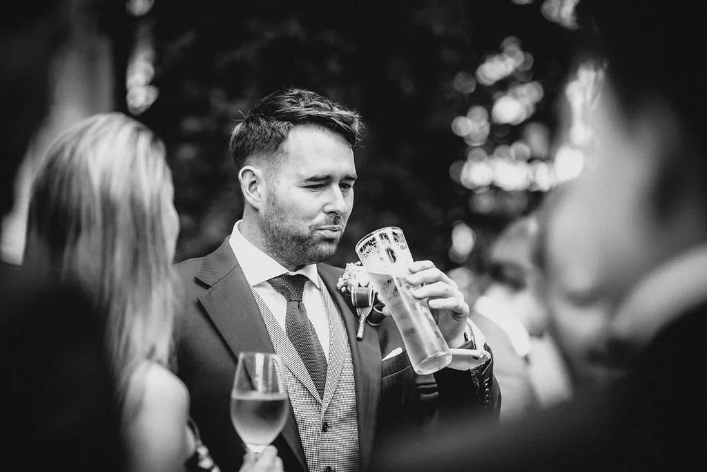 A man in a suit and tie holding a large beer mug at an outdoor social event, with other people around him.
