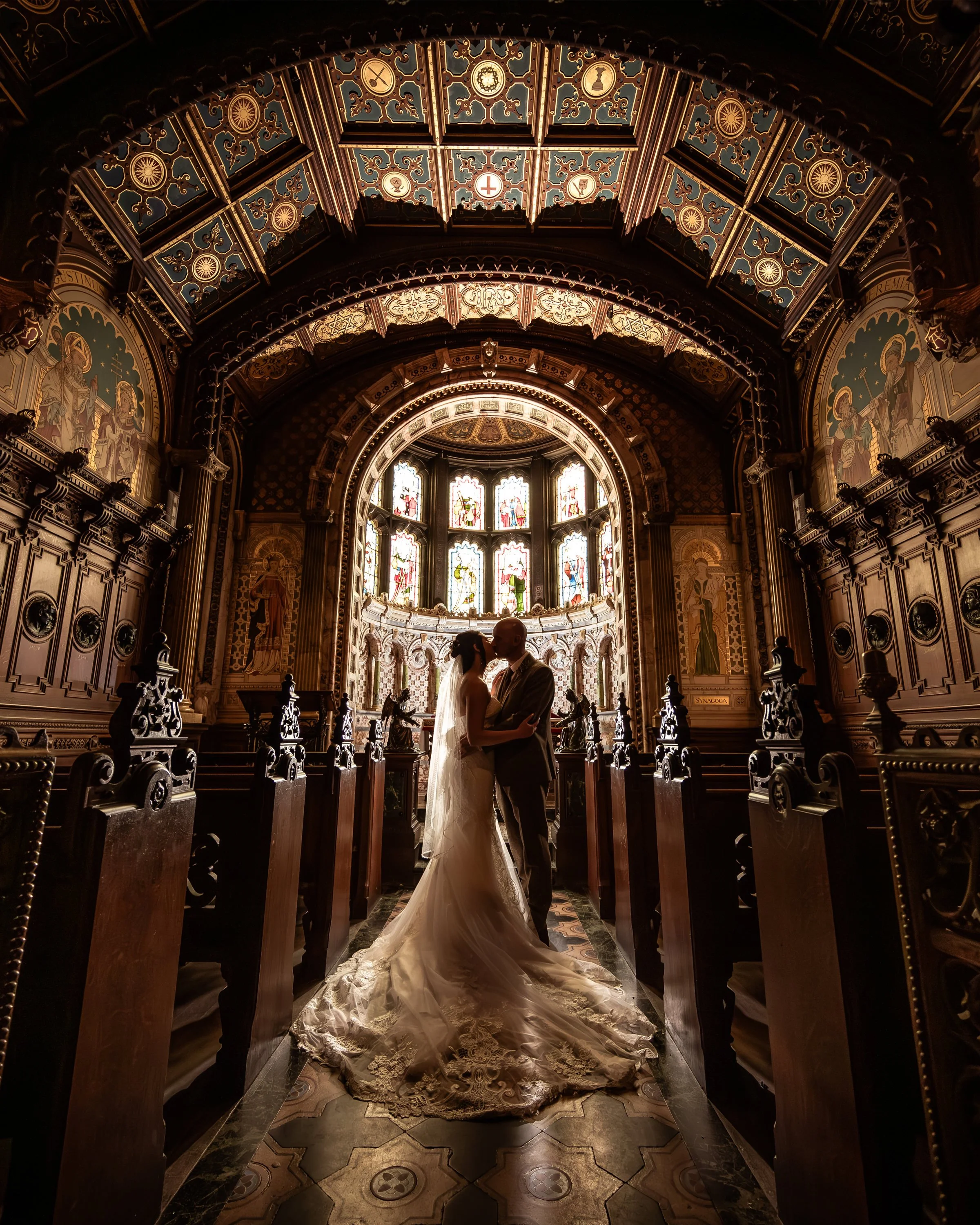 A bride and groom sharing a moment inside a church with ornate wooden pews, stained glass windows, and intricate ceiling and wall decorations.
