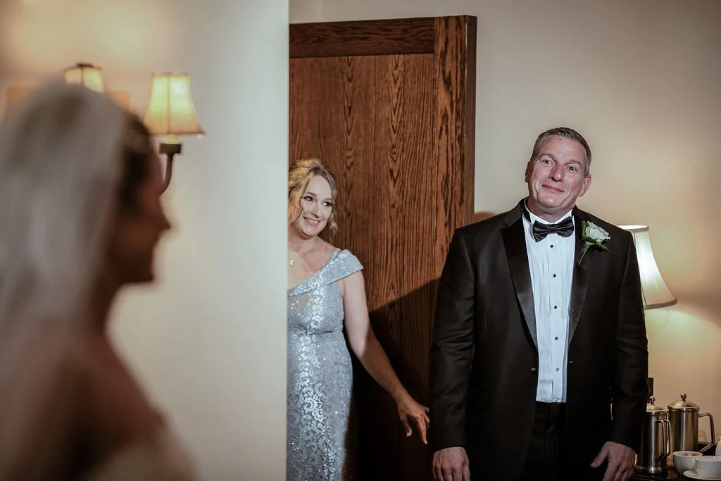 A man in a tuxedo with a bow tie and a boutonnière, smiling, is standing in a room next to a woman in a sparkly dress. Another woman, out of focus, is looking at them from the foreground.