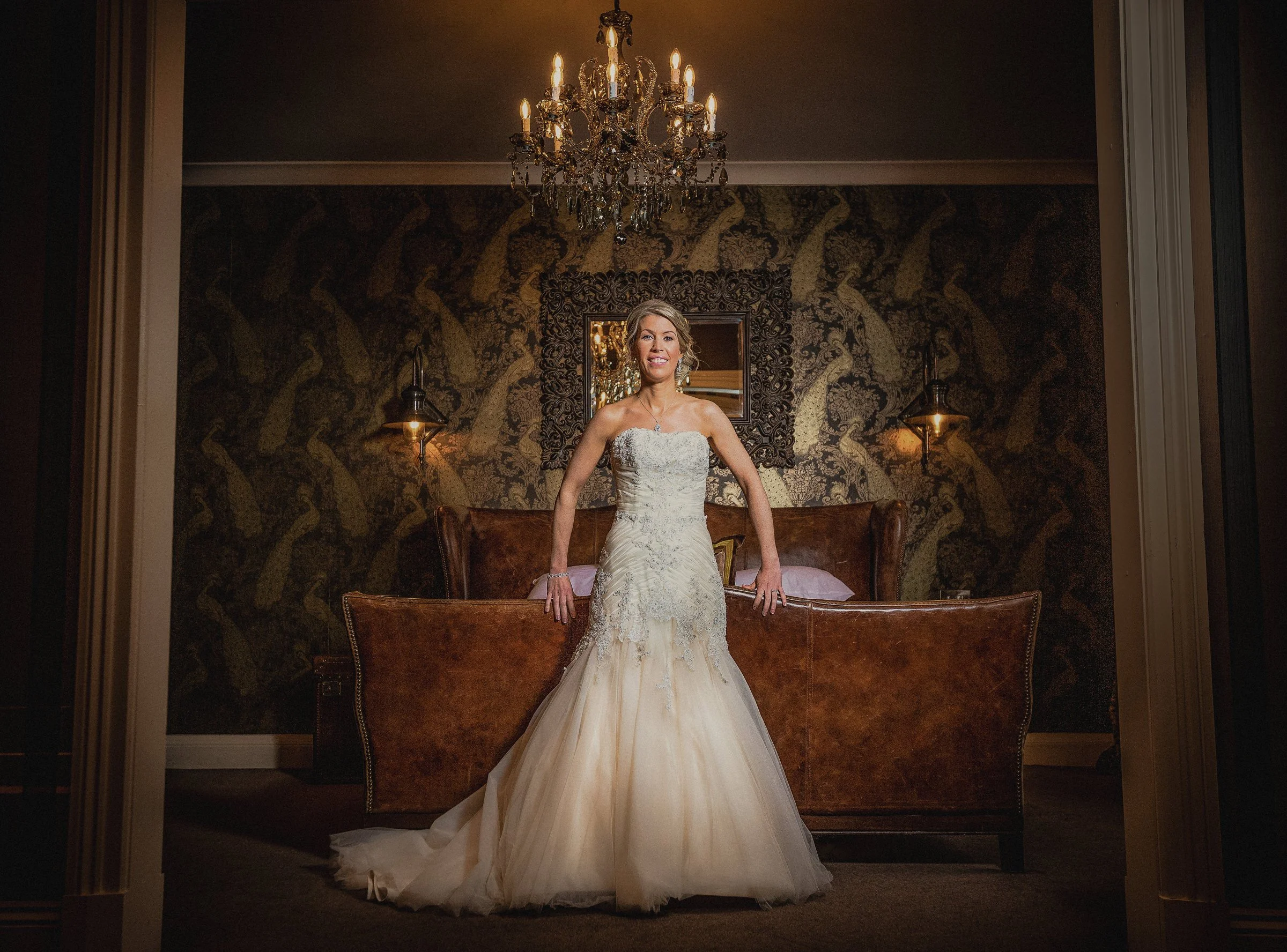 A woman in a wedding dress standing on a bed in a dimly lit room with a chandelier and wallpaper.