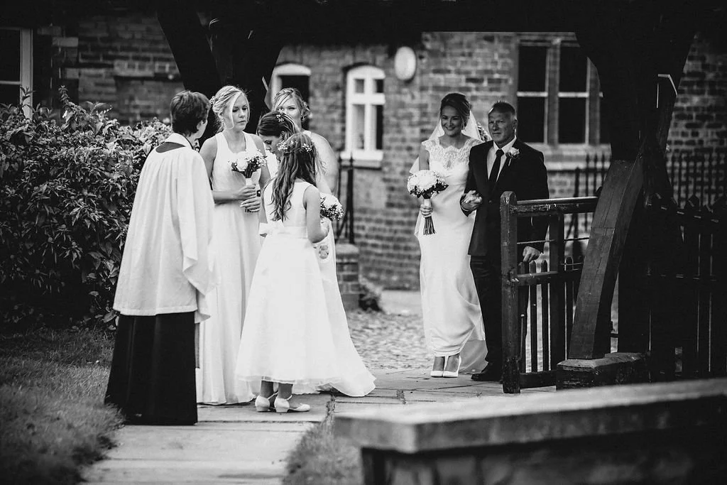 A black and white photo of a wedding ceremony with six people outside a brick building, including a bride and groom standing near a gate, and four women in dresses holding bouquets engaged in conversation.