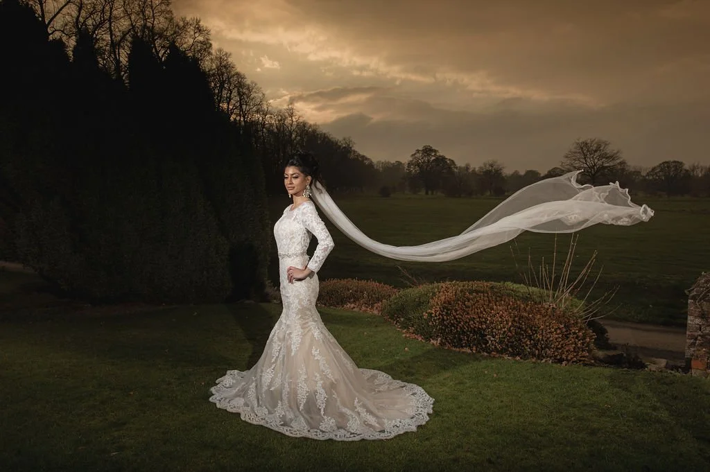 Bride in a lace wedding gown and long veil standing outdoors at dusk with a landscape of trees, bushes, and a cloudy sky in the background.