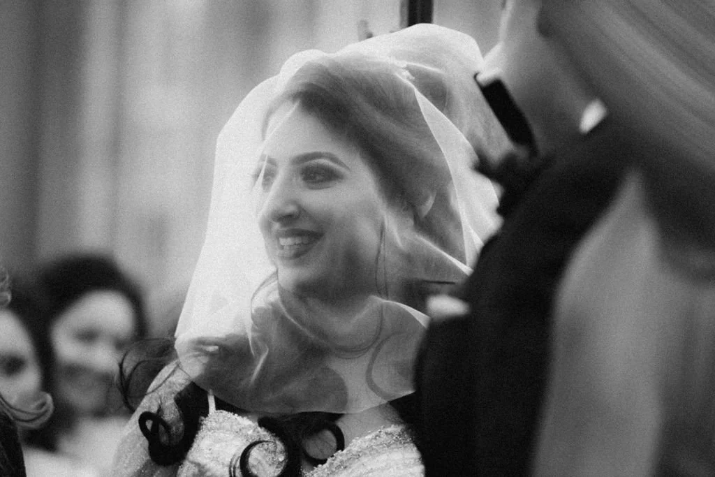 Black and white photo of a bride smiling, wearing a veil, inside a church or wedding venue. Bridesmaids and a man in a tuxedo are visible in the background.