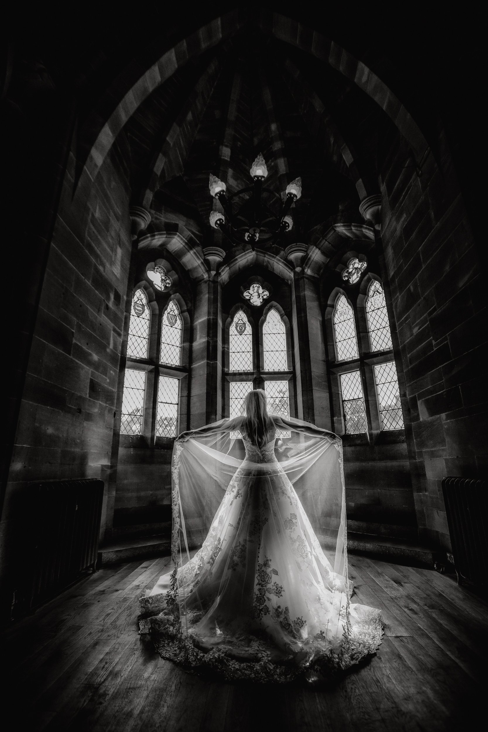 A woman in a wedding dress standing inside a dark, gothic-style church with tall stained-glass windows, holding the edges of her veil, facing away from the camera.