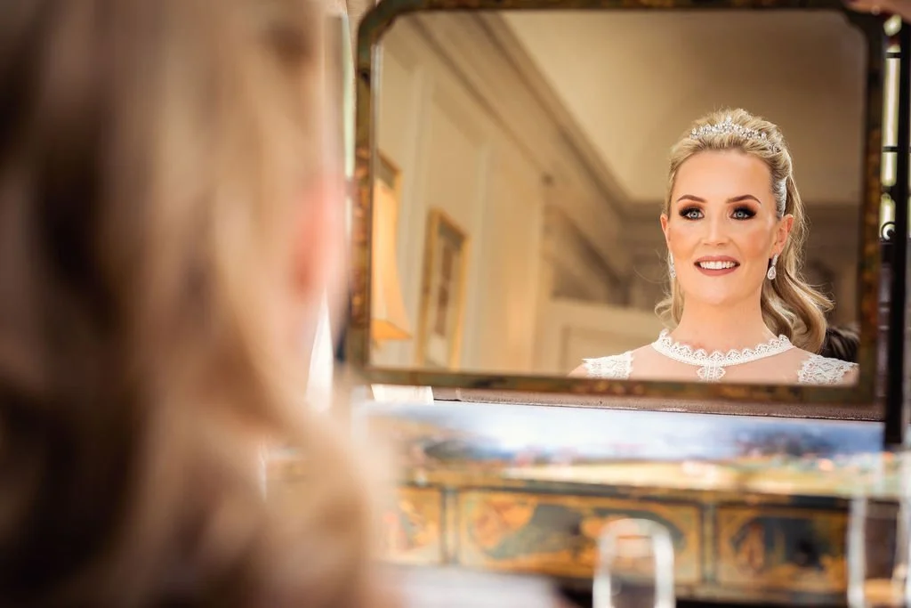 A woman with styled blonde hair and makeup looks at her reflection in an ornate mirror, wearing a white lace dress and earrings.
