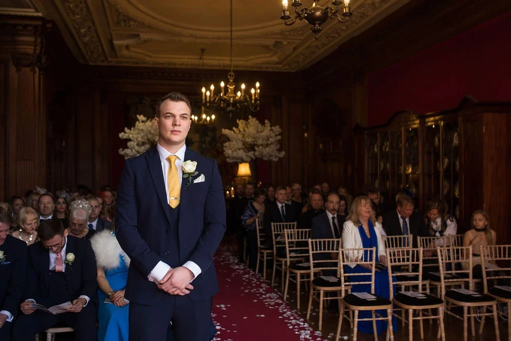 A young man in a navy suit, white shirt, and yellow tie standing among seated wedding guests in an ornately decorated room with chandeliers and floral arrangements.