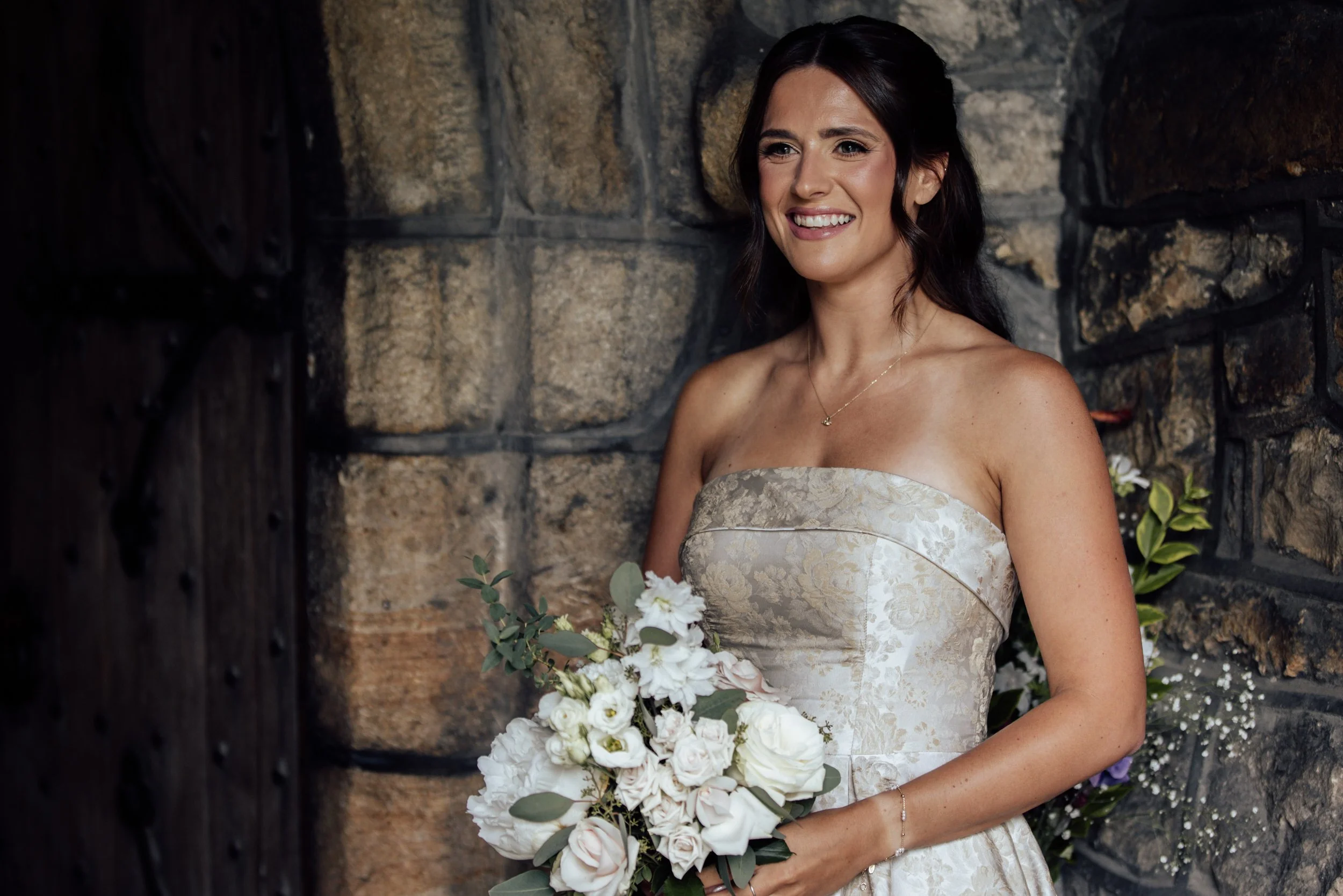 A smiling woman in a strapless wedding dress holding a bouquet of white and light pink flowers. She has dark wavy hair and wears a delicate necklace, standing in front of a stone wall.