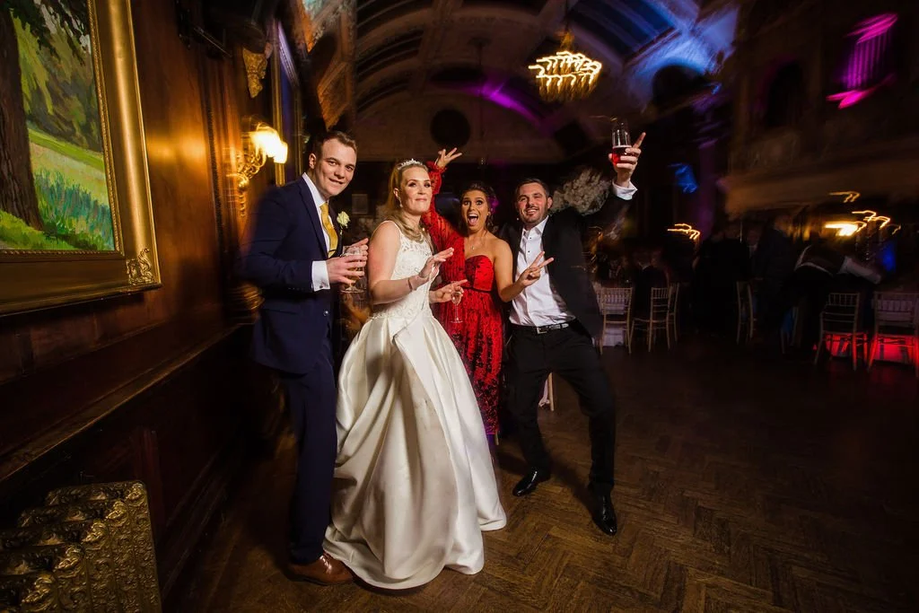 Group of four people dancing and celebrating at a wedding reception in a decorated hall, with a bride in a white gown and three guests, one holding a glass of wine, all smiling and enjoying the party.