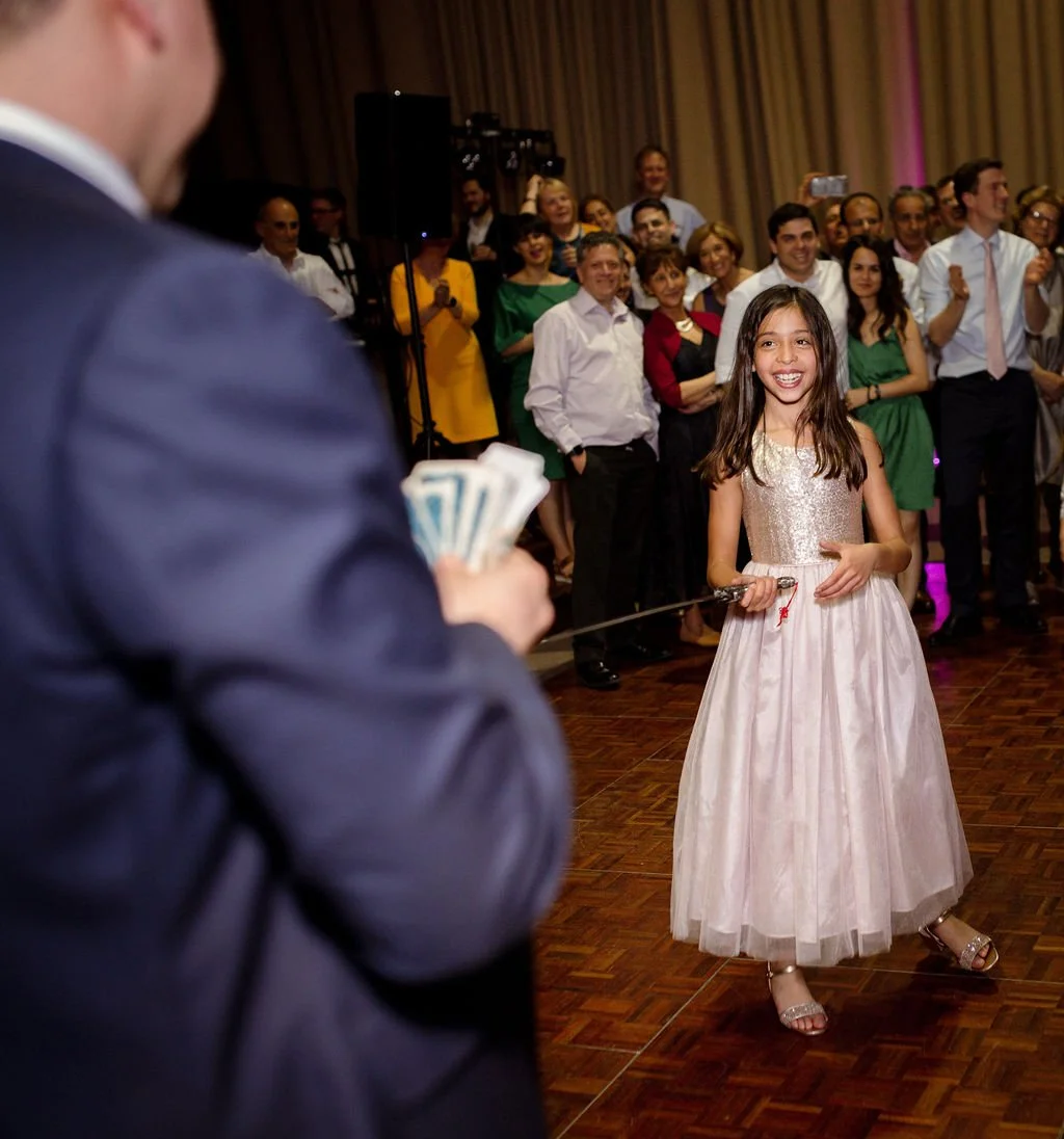 A young girl in a fancy dress is smiling and holding a fencing sword, standing on a dance floor. In the foreground, a person in a suit, partially visible, is holding a stack of money. Behind the girl, a crowd of adults is watching and smiling, some t