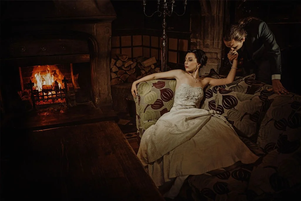A woman in a strapless dress reclining on a patterned sofa near a fireplace with a fire burning, while a man in a suit leans over to kiss her hand in a dimly lit, rustic room.