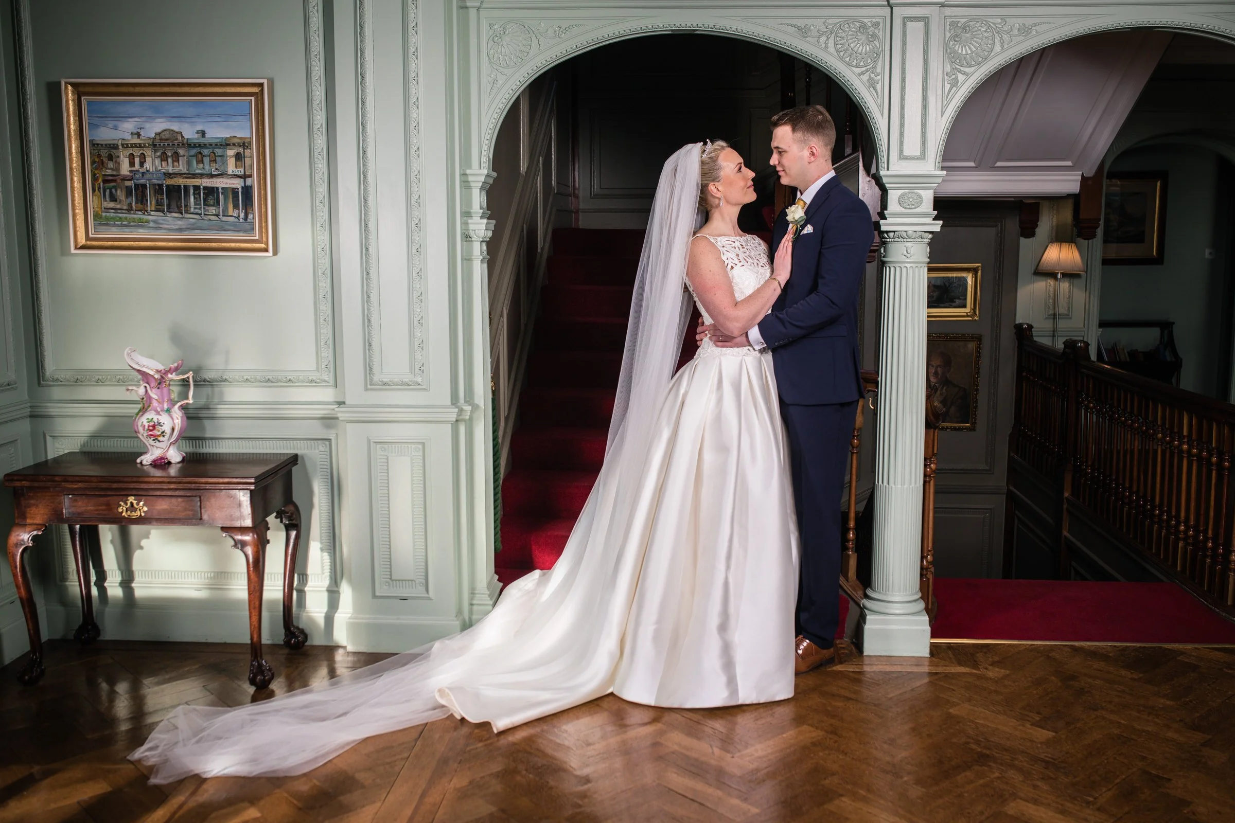 A bride and groom standing close together in an elegant, vintage-style indoor setting. The bride is wearing a white wedding gown with a long train and veil, and the groom is dressed in a navy blue suit. They are looking into each other's eyes, with the bride touching the groom's chest. The background features a staircase with a red carpet, framed artwork, a wooden side table with a pink decorative vase, and ornate wall paneling.