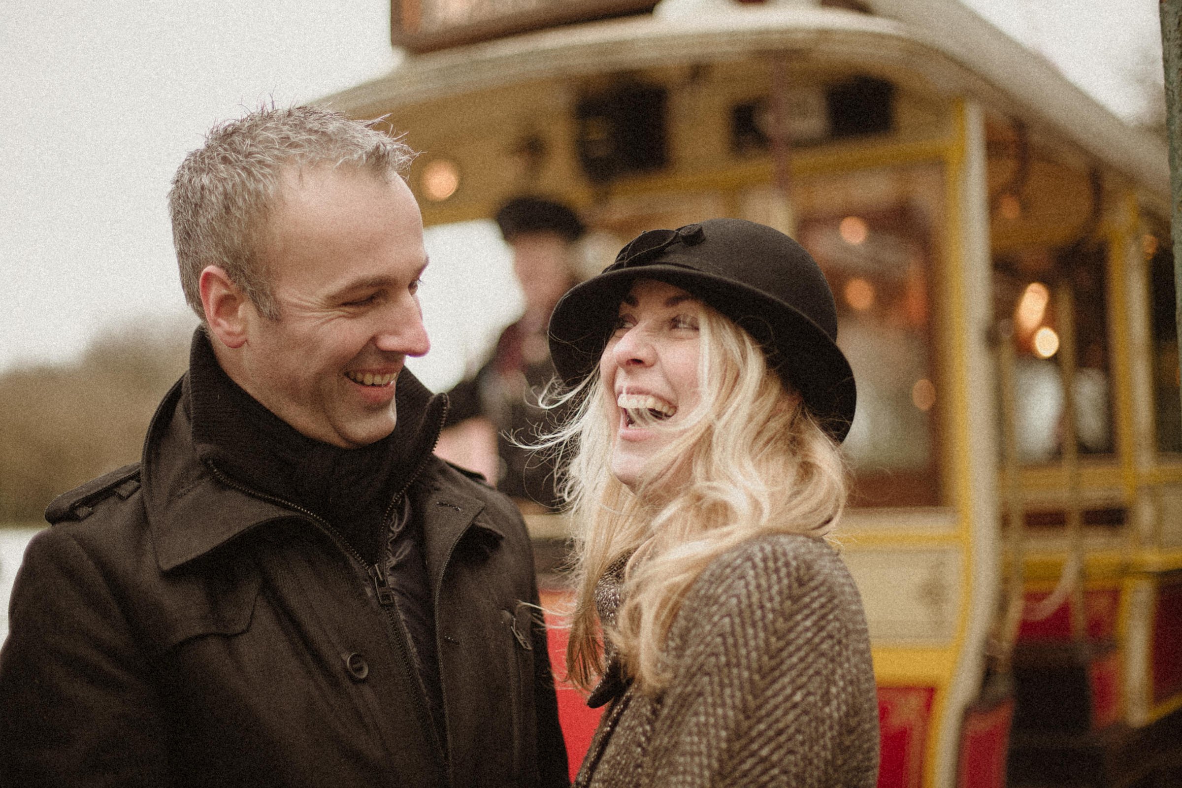 A man and a woman are smiling and laughing together outdoors with a vintage yellow and red tram in the background.