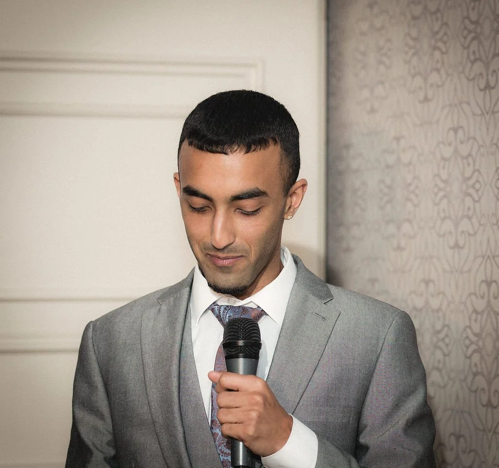 A young man in a gray suit and colorful tie holds a microphone, looking down with a slight smile.