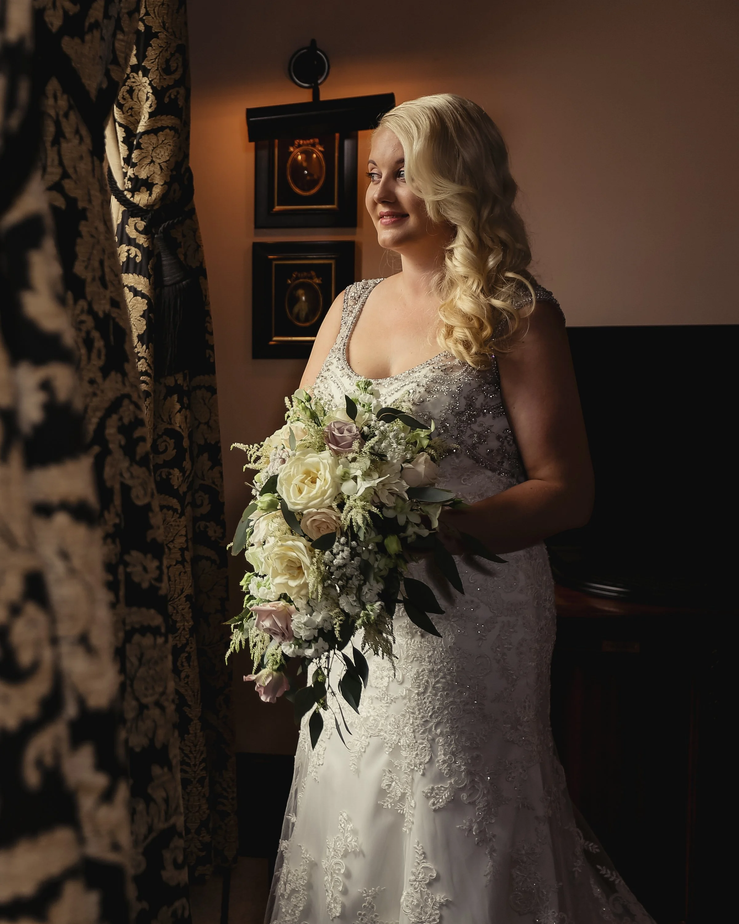 A bride with long, blonde, curled hair in a lace wedding dress holding a cascading bouquet of white and pale pink roses, standing indoors with ornate curtains and framed artwork in the background.
