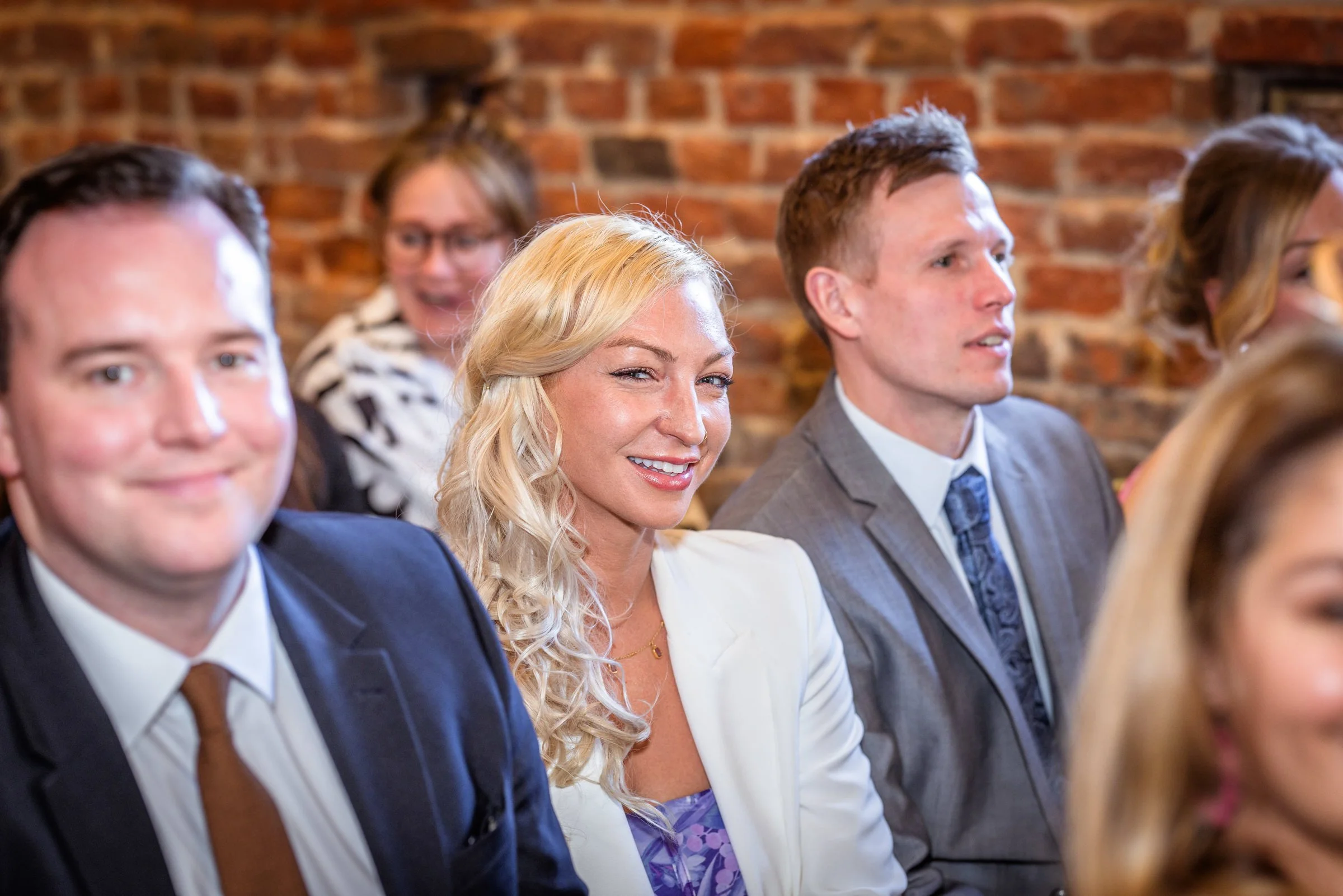 People sitting in a row at an indoor event, smiling and dressed in formal attire, with a brick wall background.